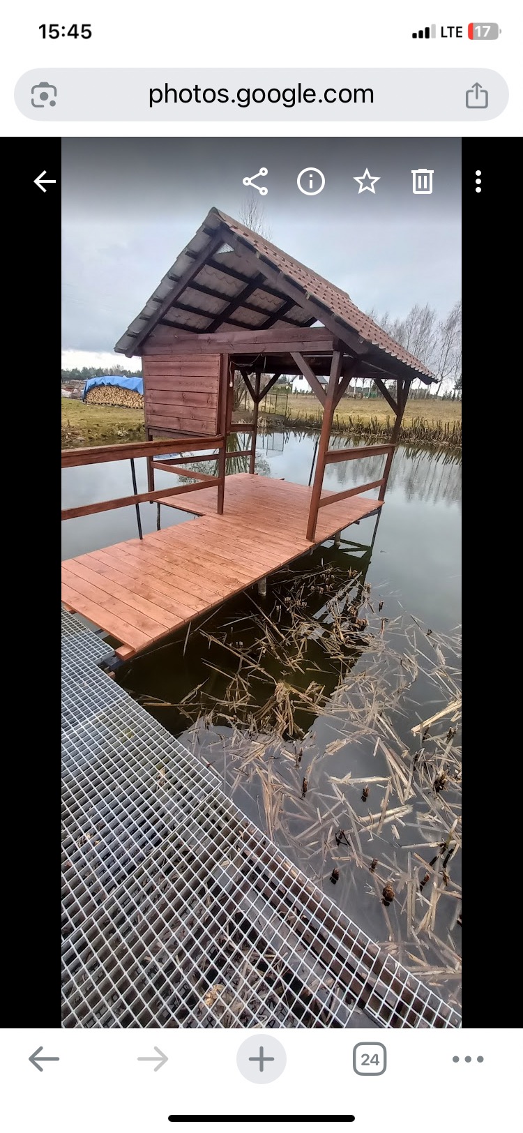 Drewniana altana z pomostem nad stawem, świeżo po impregnacji. Widoczne detale konstrukcyjne i odbicie w wodzie. W tle składowane drewno pod plandeką.