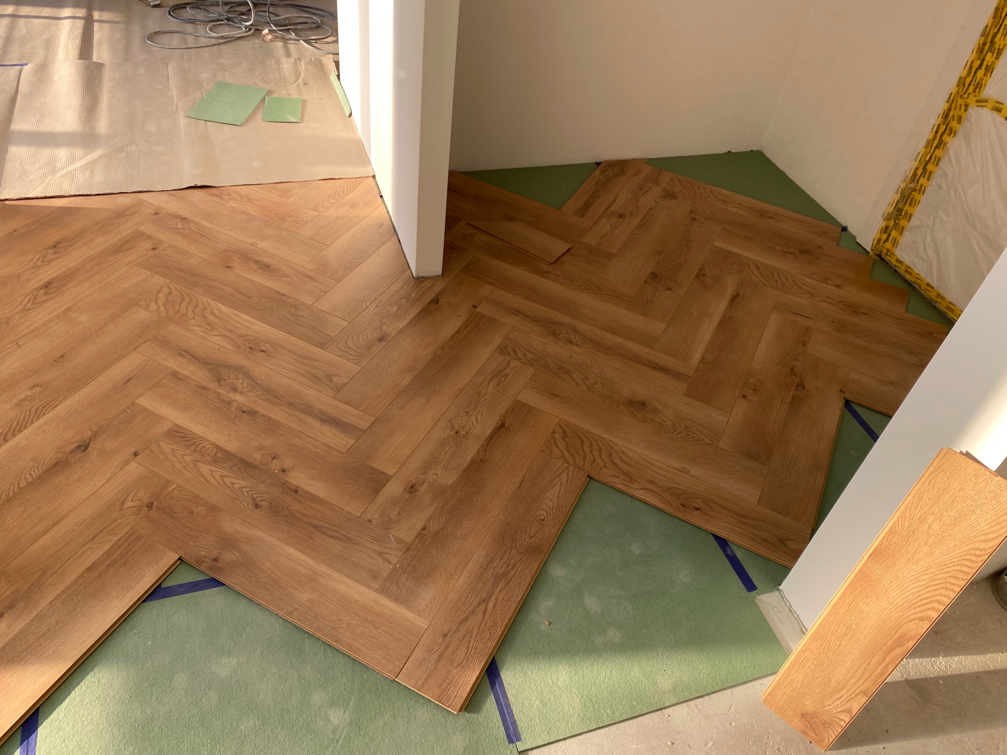 Układanie parkietu w jodełkę na zielonym podkładzie. Widoczny fragment ułożonej podłogi i przygotowane panele. Częściowo zabezpieczona ściana folią malarską.