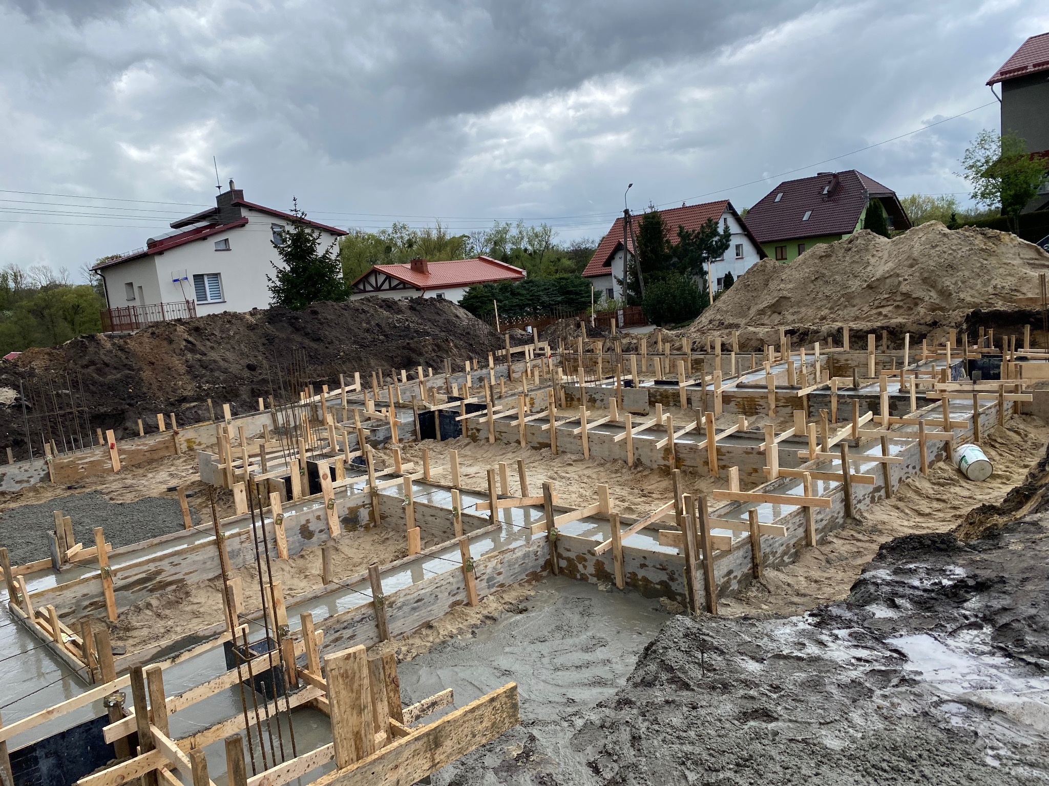 Wylane fundamenty pod budowę domu jednorodzinnego. Widoczne drewniane szalunki wypełnione betonem, zbrojenie, piasek i ziemia. W tle zabudowa jednorodzinna i pochmurne niebo.