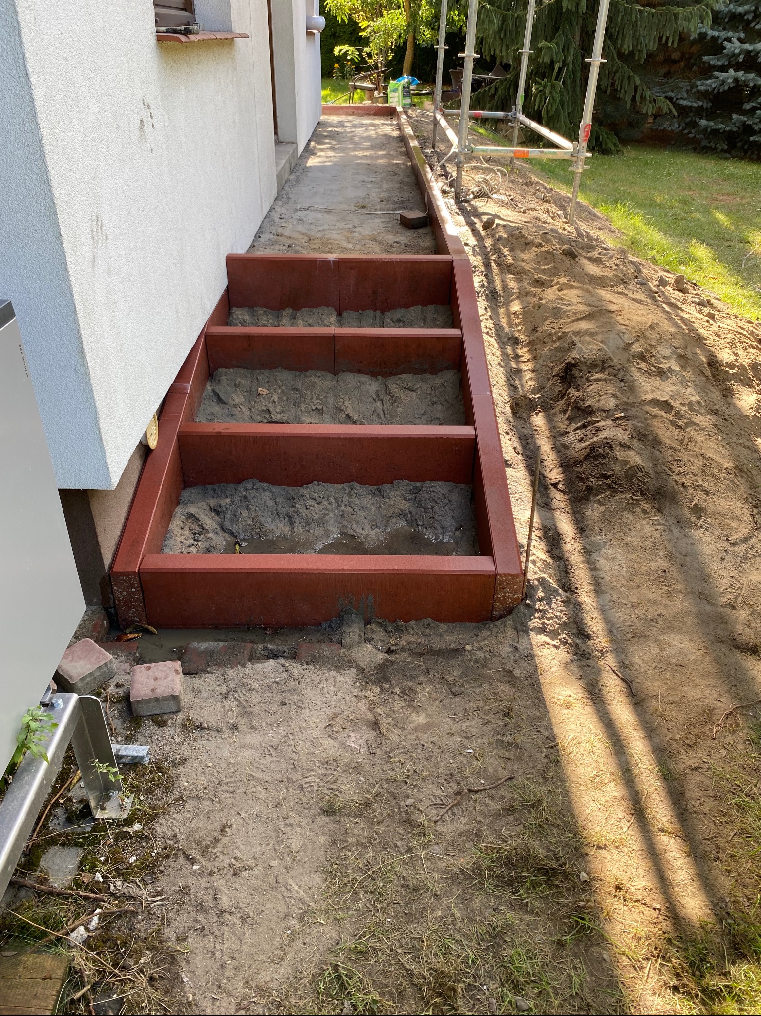 Konstrukcja schodów zewnętrznych z czerwonej kostki, częściowo wypełniona ziemią, przylegająca do białej ściany budynku. Widoczne rusztowanie i teren budowy.