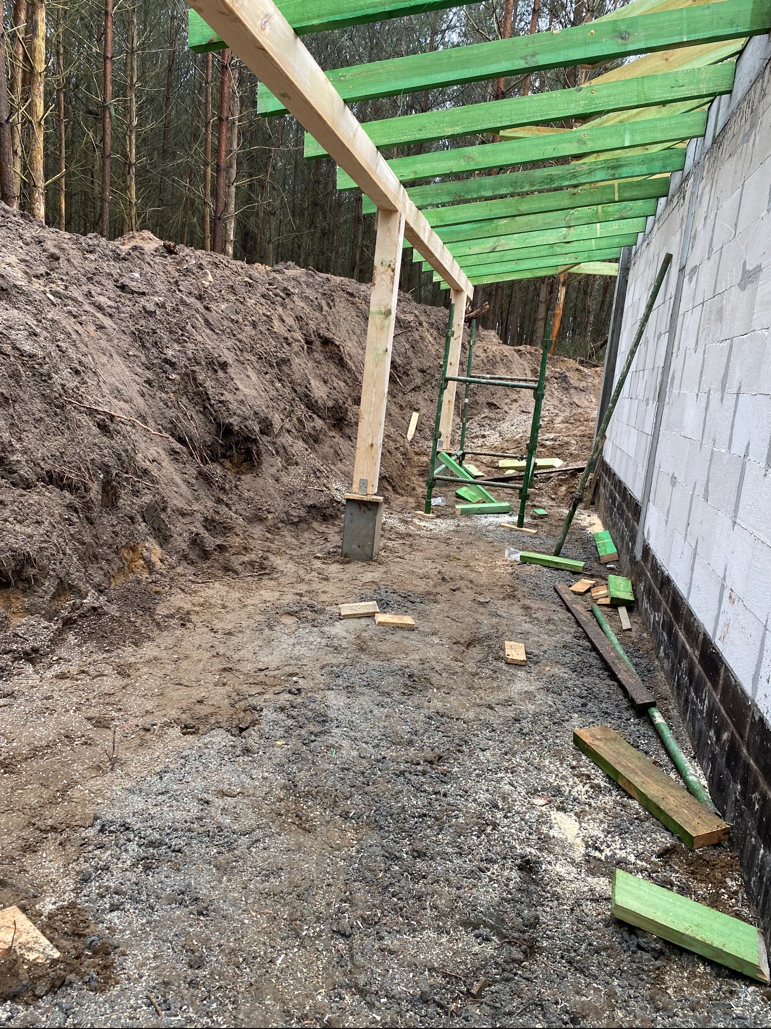 Wznoszenie drewnianej konstrukcji z zielonymi elementami przy ścianie z bloczków betonowych. Widoczne rusztowanie i ziemne podłoże. Prace budowlane w toku.