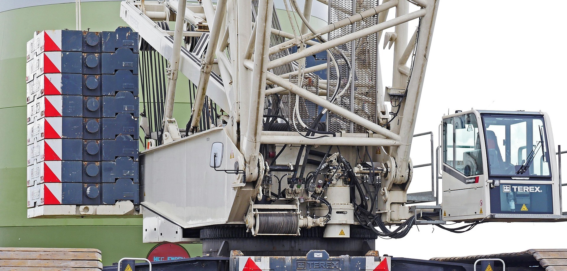 Fragment żurawia wieżowego Terex z kabiną operatora i przeciwwagą. Widoczne elementy konstrukcyjne, liny i hydraulika. Ujęcie z dołu, jasne tło.