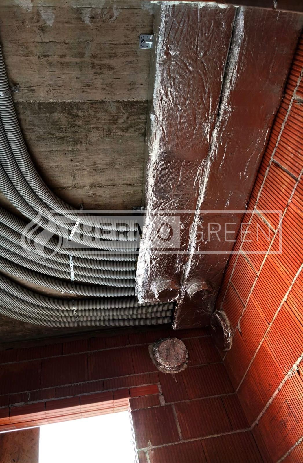 Montaż instalacji wentylacyjnej: izolowane kanały wentylacyjne z folii aluminiowej i rury karbowane przymocowane do sufitu w surowym wnętrzu z ceglanymi ścianami.