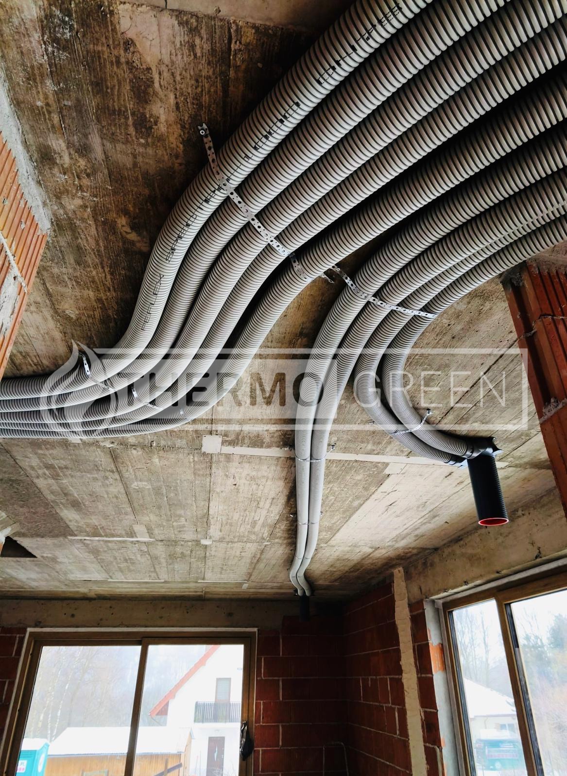 Szereg szarych, elastycznych rur wentylacyjnych przymocowanych do surowego betonowego sufitu w budynku w trakcie budowy. Widoczne okna i fragmenty ścian z cegły.