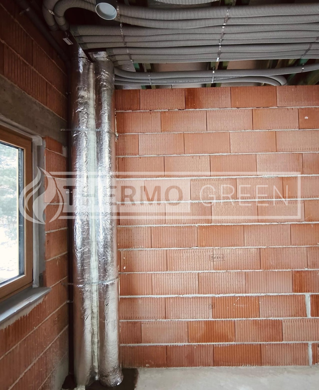 Dwie pionowe rury w srebrnej izolacji, zamocowane przy ceglanej ścianie w surowym stanie, z widocznymi przewodami pod sufitem. Instalacja w trakcie montażu.