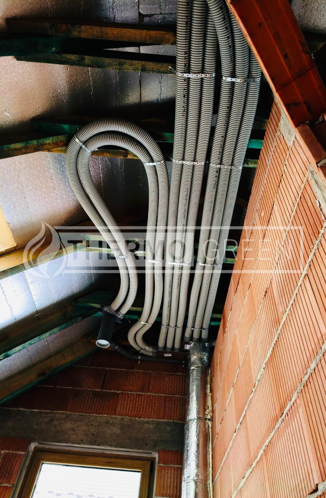 Szare, elastyczne rury wentylacyjne zamocowane do sufitu poddasza z widoczną izolacją i fragmentem murowanej ściany. Instalacja w trakcie montażu.