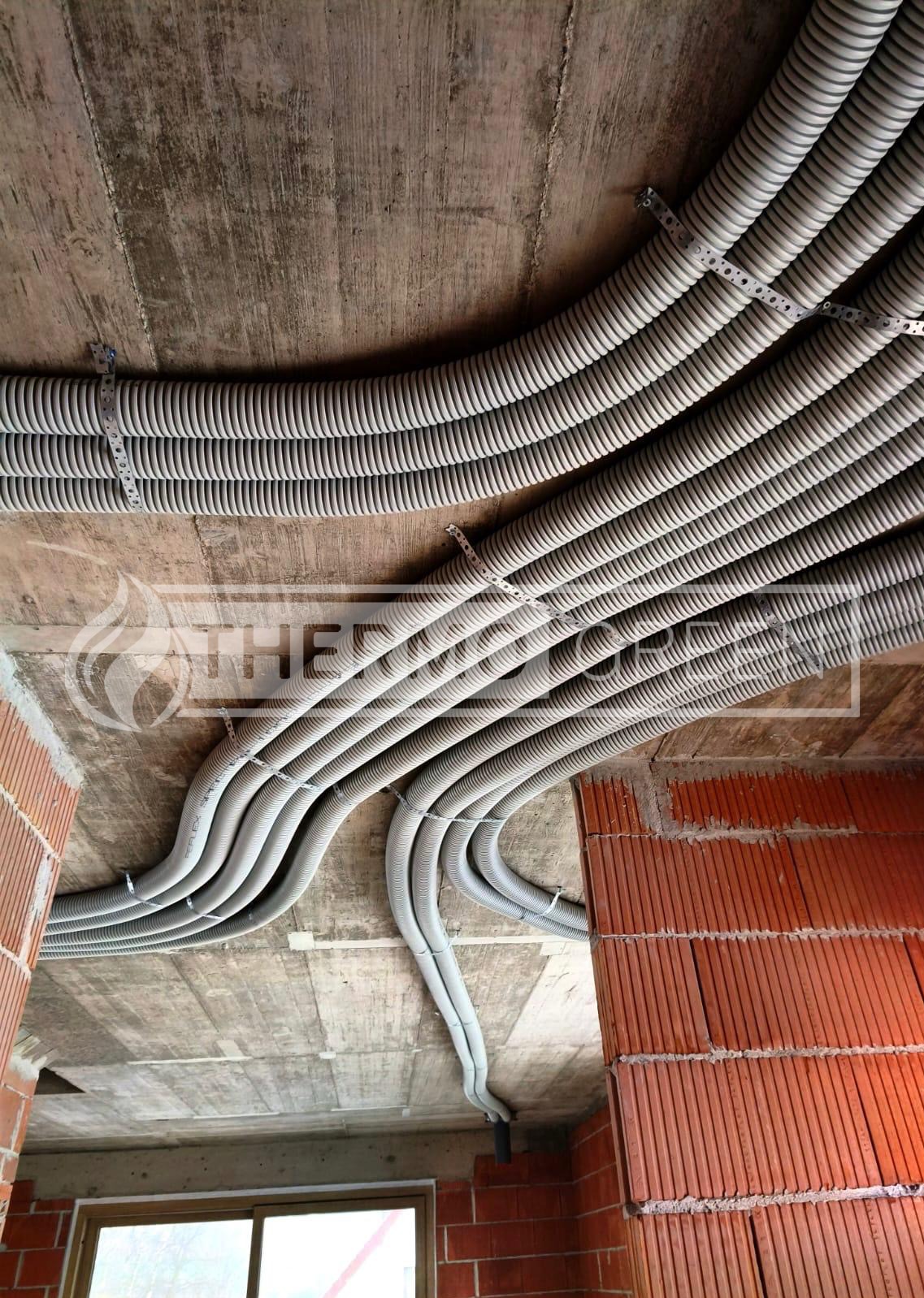 Szare, karbowane rury wentylacyjne przymocowane do surowego betonowego stropu w budynku w trakcie budowy, widoczne ceglane ściany i okno.