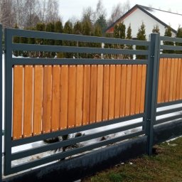 Alfa f.p.h.u - Nowoczesne ogrodzenie panelowe z drewnianymi wypełnieniami, szare słupki i poziome elementy, widoczne śruby mocujące, trawa i śnieg w tle.