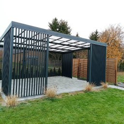 Alfa f.p.h.u - Nowoczesna, grafitowa pergola metalowa z przezroczystym zadaszeniem na tle zielonego trawnika i ogrodzenia. Minimalistyczny design, pionowe lamele, kostka brukowa wewnątrz.