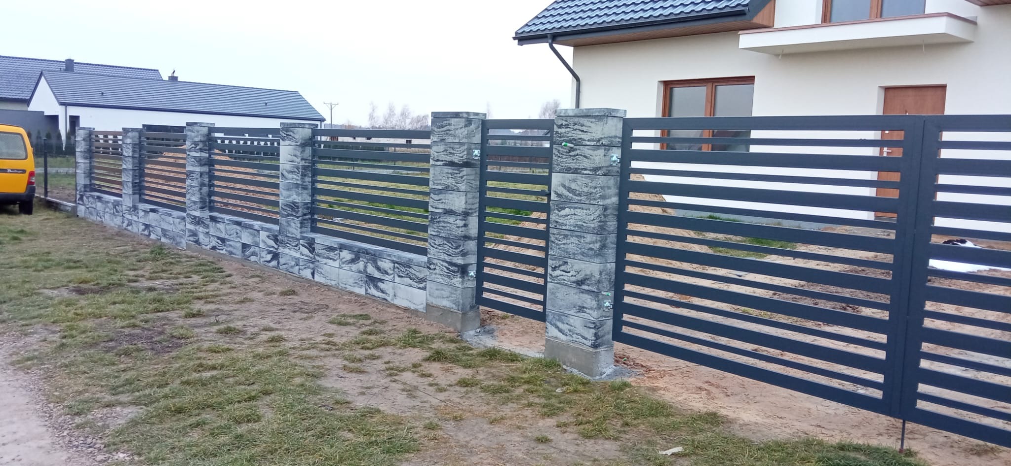 Nowoczesne ogrodzenie panelowe z elementami imitującymi kamień, szare, poziome panele, widok z zewnątrz posesji, dom w tle.