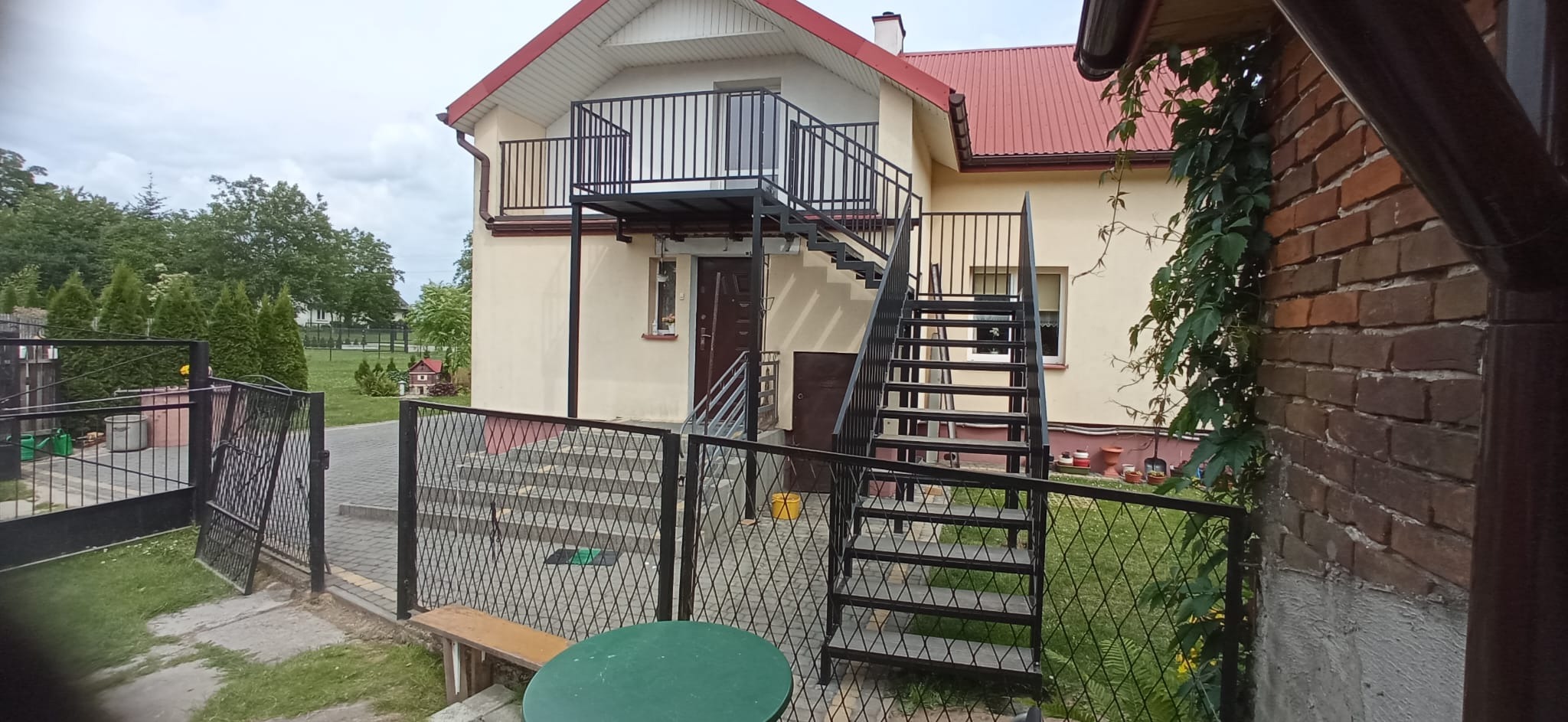 Zewnętrzne schody metalowe z balustradą prowadzące na balkon domu jednorodzinnego. Widoczne ogrodzenie z siatki i fragment zieleni.