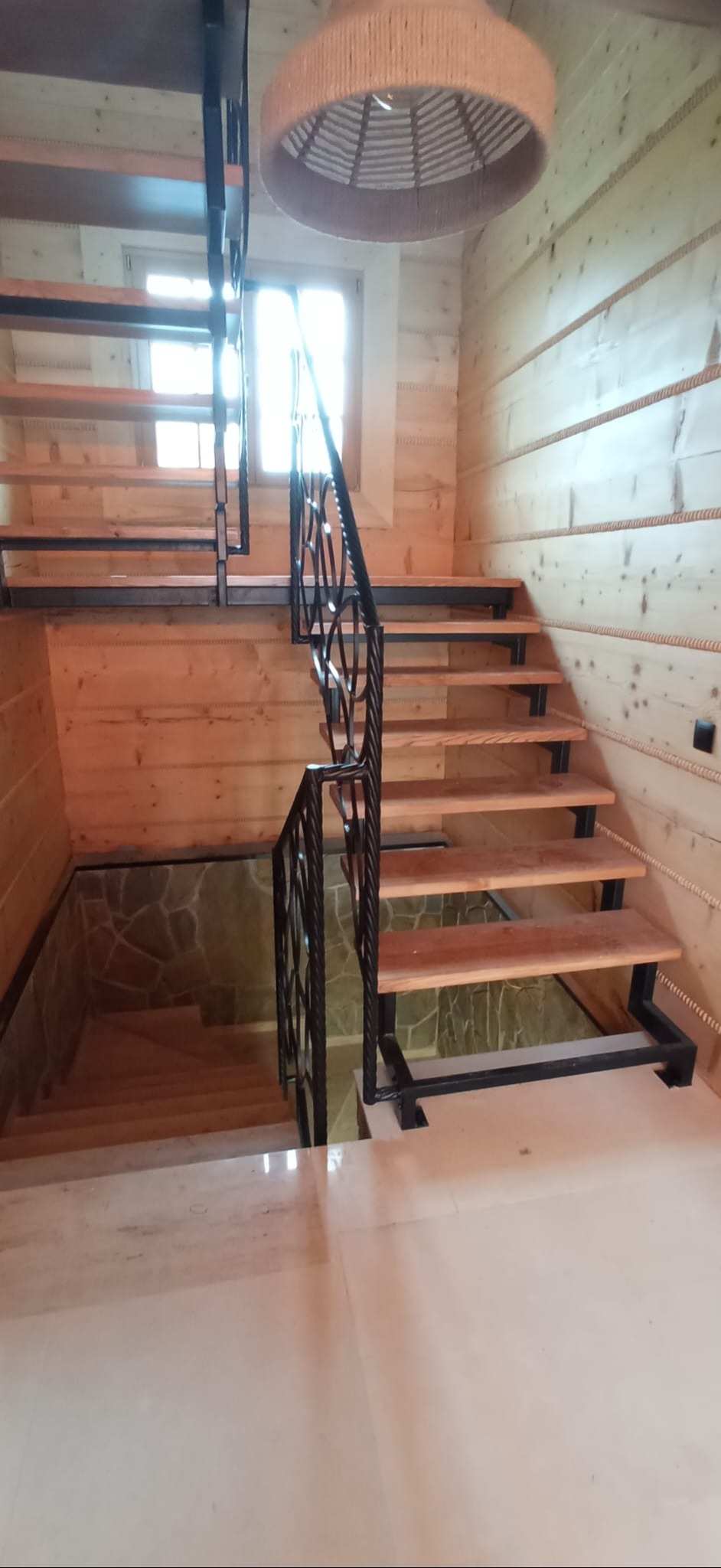 Drewniane schody z metalową, zdobioną balustradą w domu z boazerią. Widoczne stopnie i fragment podłogi. U góry lampa z abażurem. Jasne wnętrze.