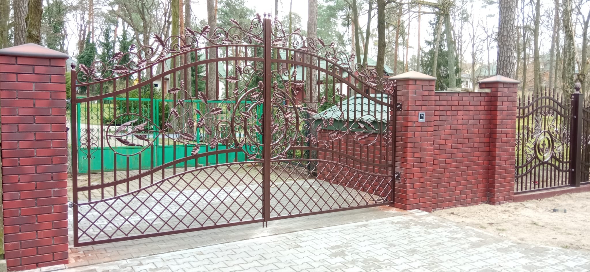Kuta brama wjazdowa z dekoracyjnymi elementami roślinnymi, osadzona na murowanych słupkach z czerwonej cegły. W tle widoczny fragment ogrodzenia i zieleń.