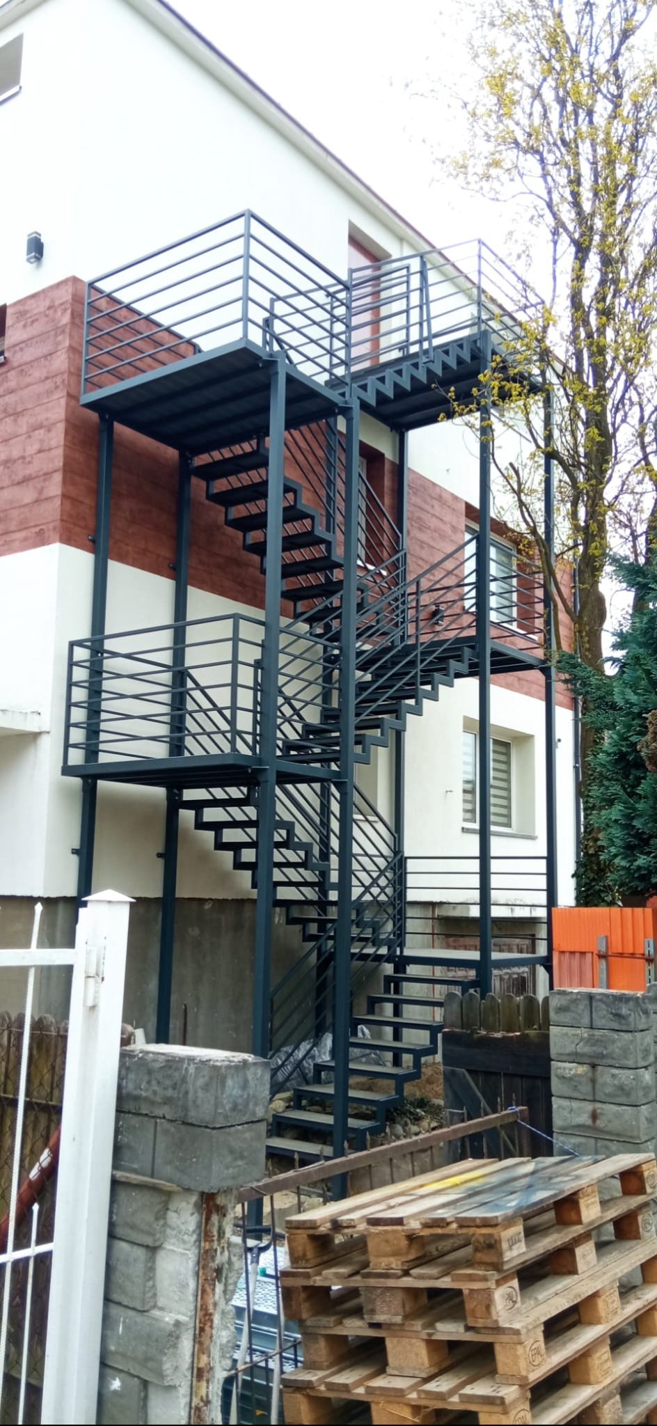 Zewnętrzne schody metalowe z balustradami, prowadzące na wyższe kondygnacje budynku. Konstrukcja w kolorze grafitowym, widok z dołu, palety drewniane w tle.