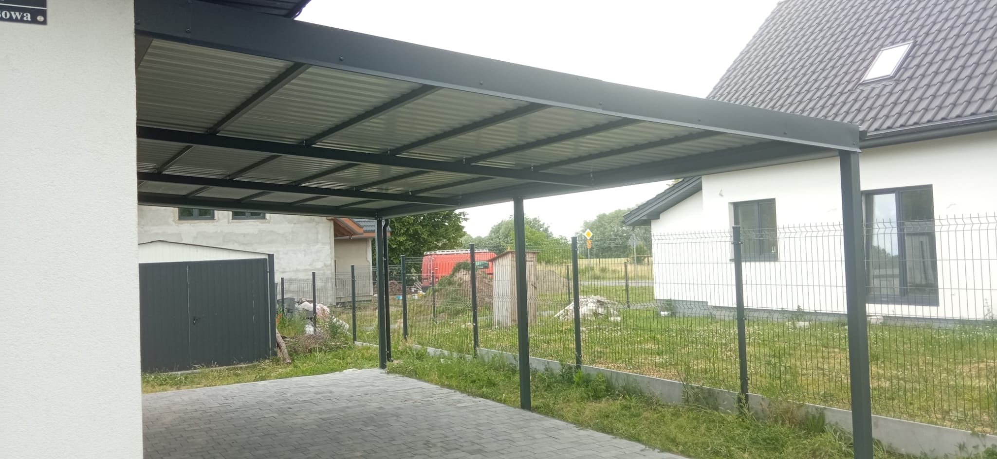 Nowoczesna, grafitowa pergola metalowa z falistym zadaszeniem, wsparta na słupach, na tle kostki brukowej i ogrodzenia panelowego. Widok na dom i sąsiednią posesję.