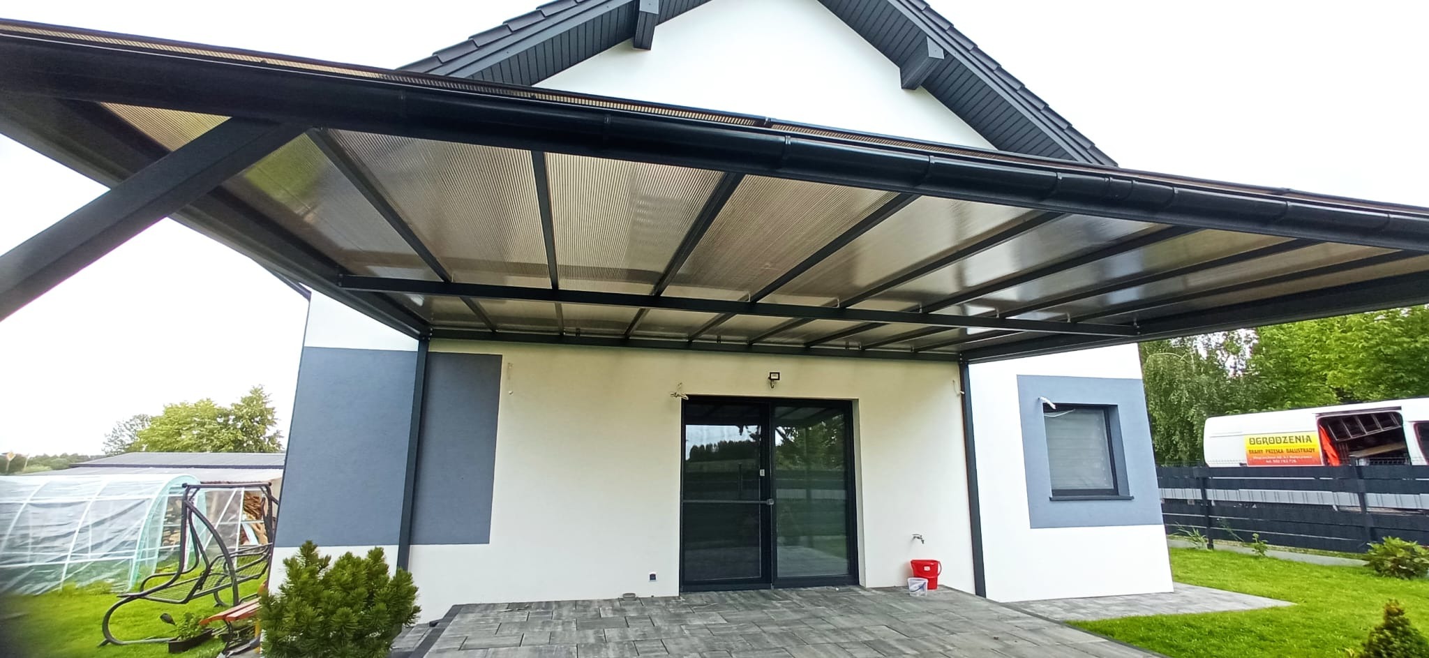 Nowoczesna, czarna pergola metalowa z przezroczystym zadaszeniem, przylegająca do ściany domu z szarą kostką brukową. Widoczny fragment ogrodu i samochód dostawczy.