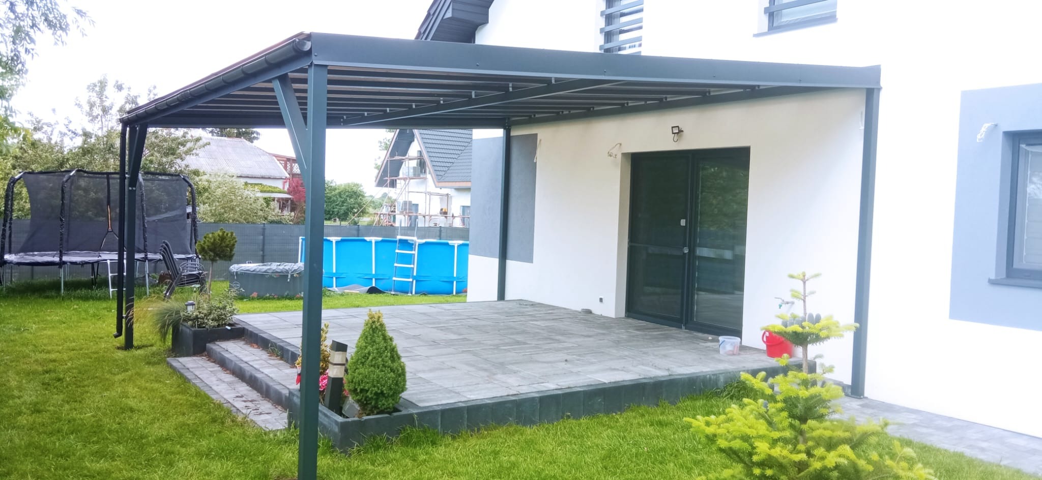 Nowoczesna, grafitowa pergola metalowa przylegająca do białego domu z szarymi akcentami. Widoczny taras z kostki brukowej, trawnik, trampolina i basen w tle.