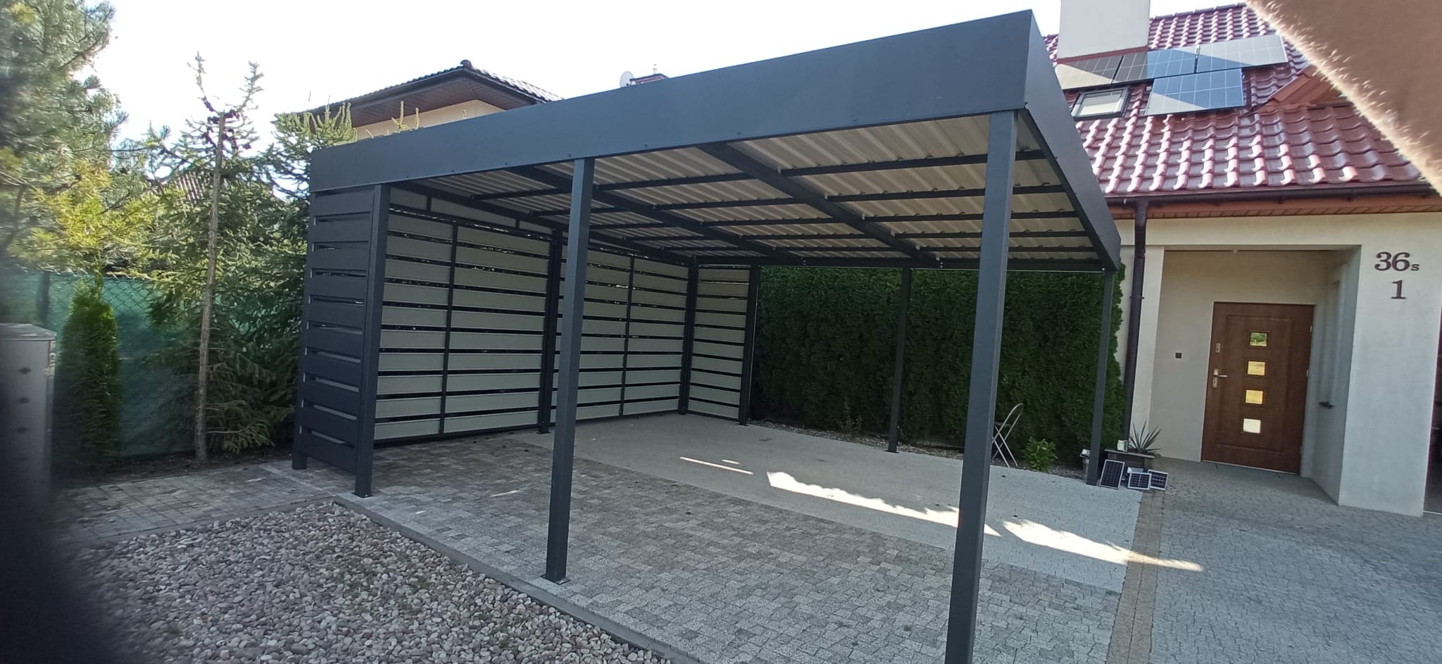 Nowoczesna, grafitowa pergola metalowa z ażurowymi ścianami bocznymi, na tle domu z panelami słonecznymi i kostki brukowej. Widok z zewnątrz, w słoneczny dzień.