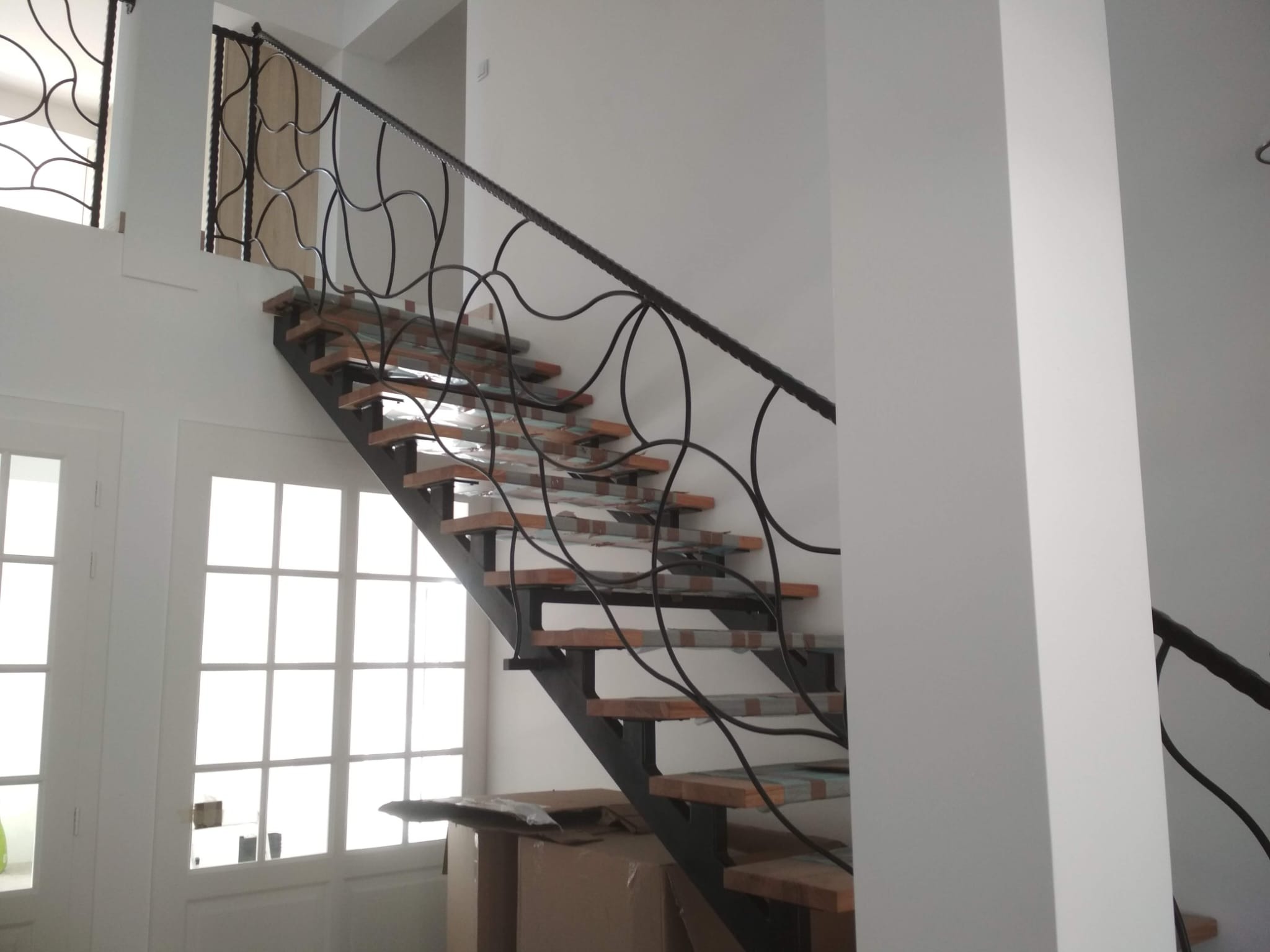 Nowoczesne schody metalowe z drewnianymi stopniami i ozdobną, kręconą balustradą, częściowo zabezpieczone folią, w jasnym wnętrzu domu.