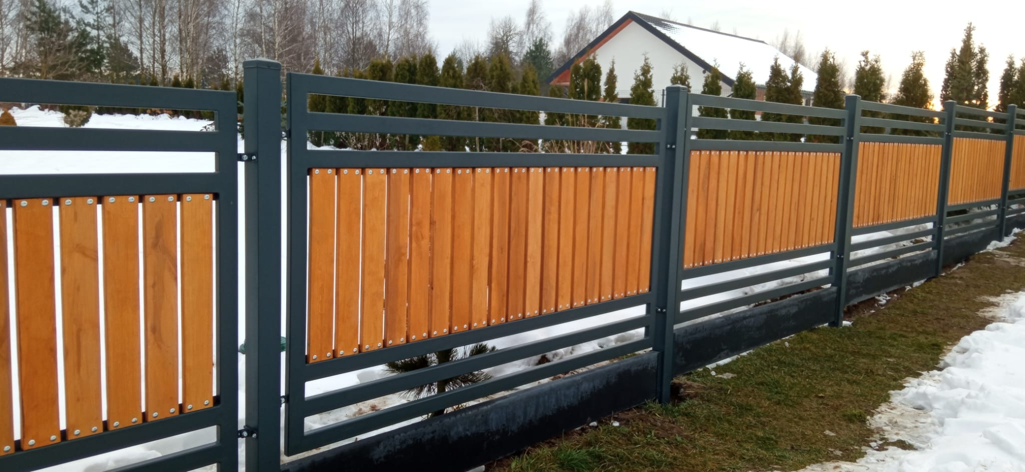 Nowoczesne ogrodzenie panelowe z drewnianymi wypełnieniami, szare słupki i poziome elementy, widoczne śruby mocujące, trawa i śnieg w tle.
