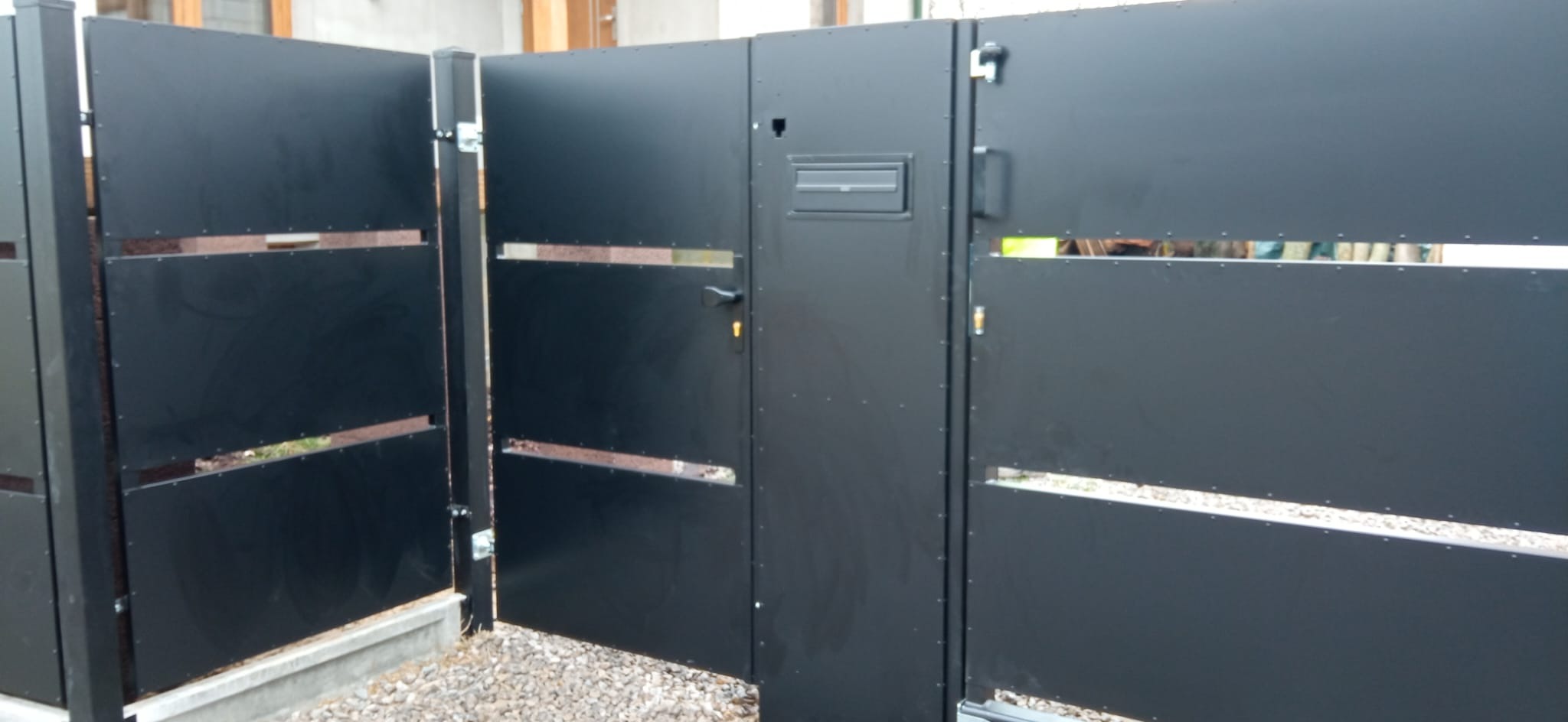 Nowoczesna, czarna brama wjazdowa z furtką i skrzynką na listy, z poziomymi prześwitami, zamontowana na posesji z szarą nawierzchnią żwirową.