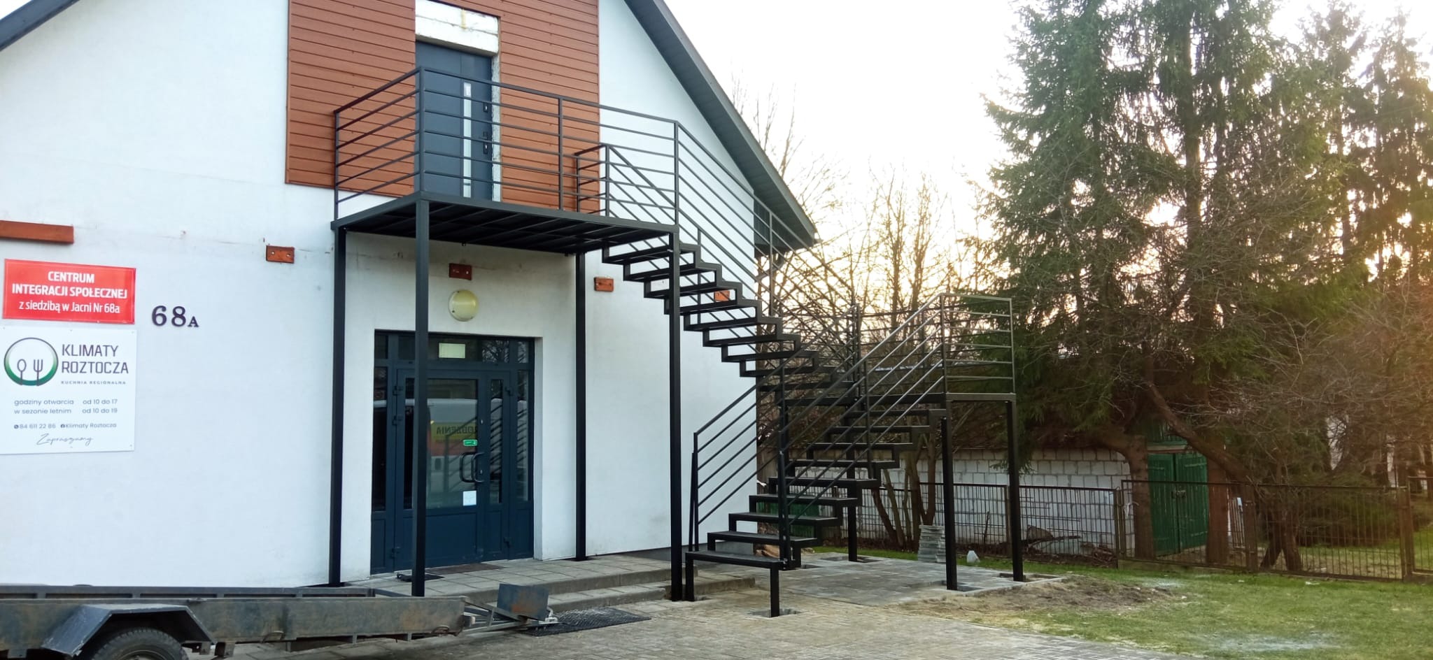 Zewnętrzne schody metalowe z platformą prowadzące do drzwi na piętrze budynku z białą elewacją i brązowymi panelami. Widoczne drzewa w tle.