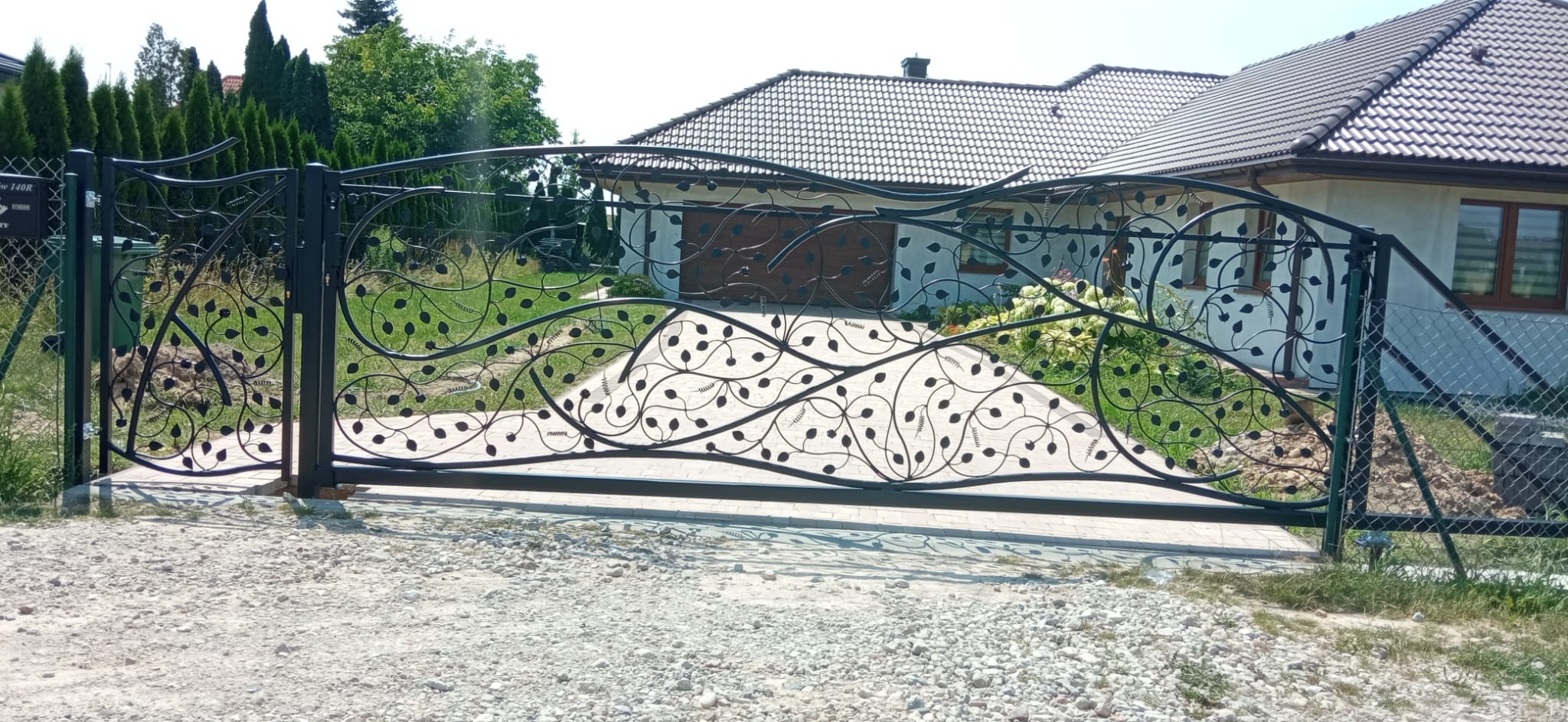Czarna, kuta brama wjazdowa z motywem liści na tle domu z brązowym dachem i zieleni. Brama zamontowana na posesji, widok z zewnątrz.