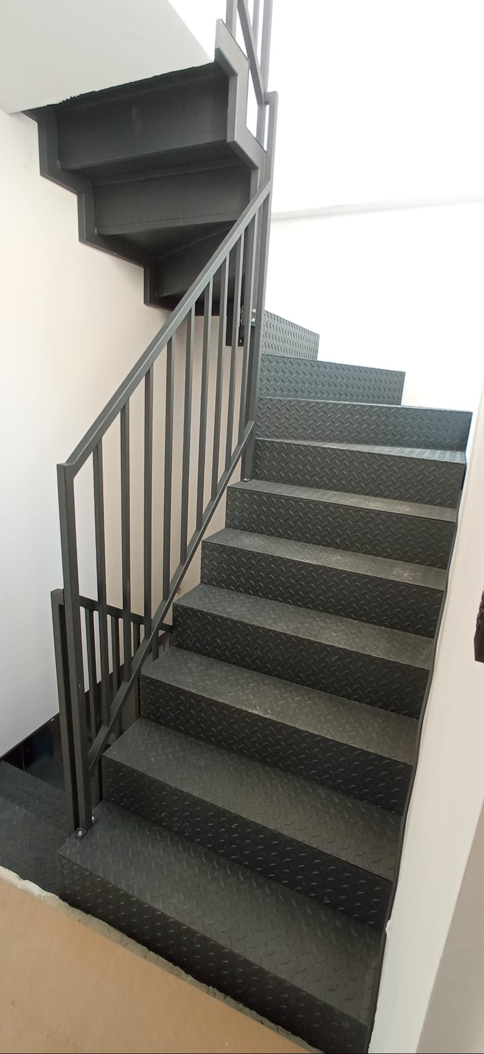 Metalowe schody z antypoślizgową fakturą i prostą balustradą w nowoczesnym wnętrzu. Widok z dołu, podkreślający solidność konstrukcji i minimalistyczny design.