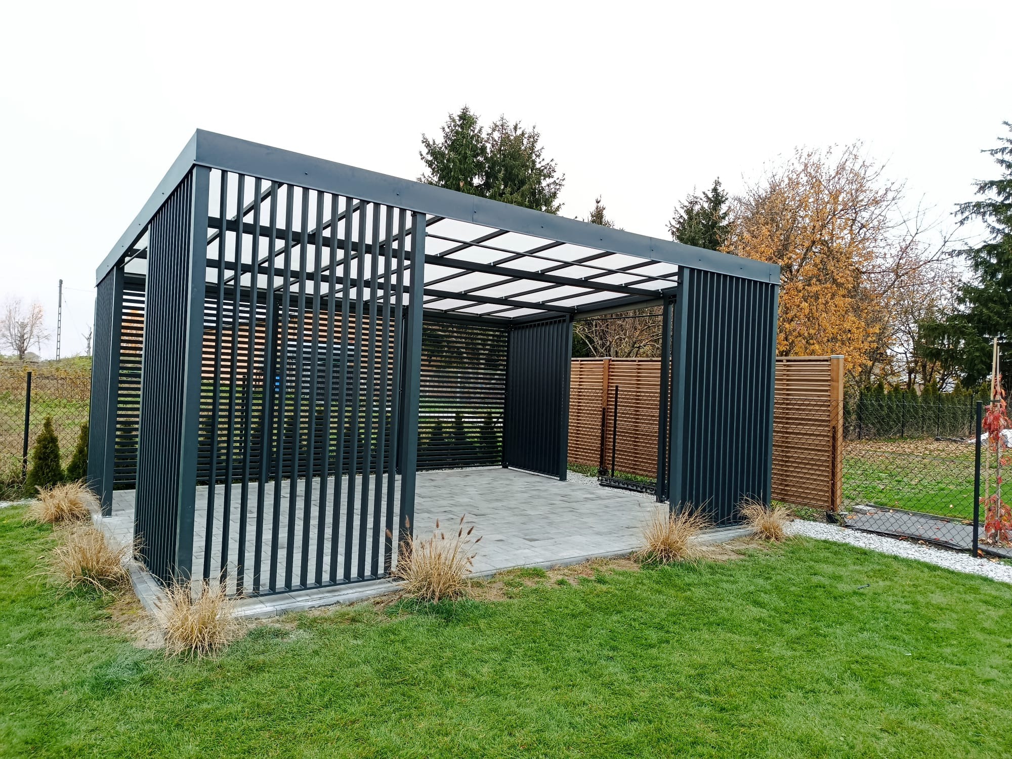 Nowoczesna, grafitowa pergola metalowa z przezroczystym zadaszeniem na tle zielonego trawnika i ogrodzenia. Minimalistyczny design, pionowe lamele, kostka brukowa wewnątrz.