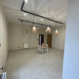 Jasne wnętrze po remoncie z szarą podłogą z płytek, białymi ścianami i drewnianymi lampami wiszącymi. Widoczny stół z krzesłami i grzejnik.
