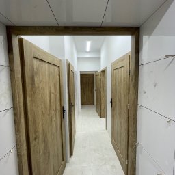 PROFI BAU - Wykończenie łazienki w Raszkowie: widok na korytarz z drewnianymi drzwiami i białymi płytkami, z widocznymi elementami montażowymi na ścianach.
