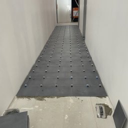 Układanie ciemnoszarych płytek podłogowych w korytarzu, z użyciem klipsów poziomujących. Widoczne narzędzia glazurnicze i zapas płytek. Surowe ściany w tle.