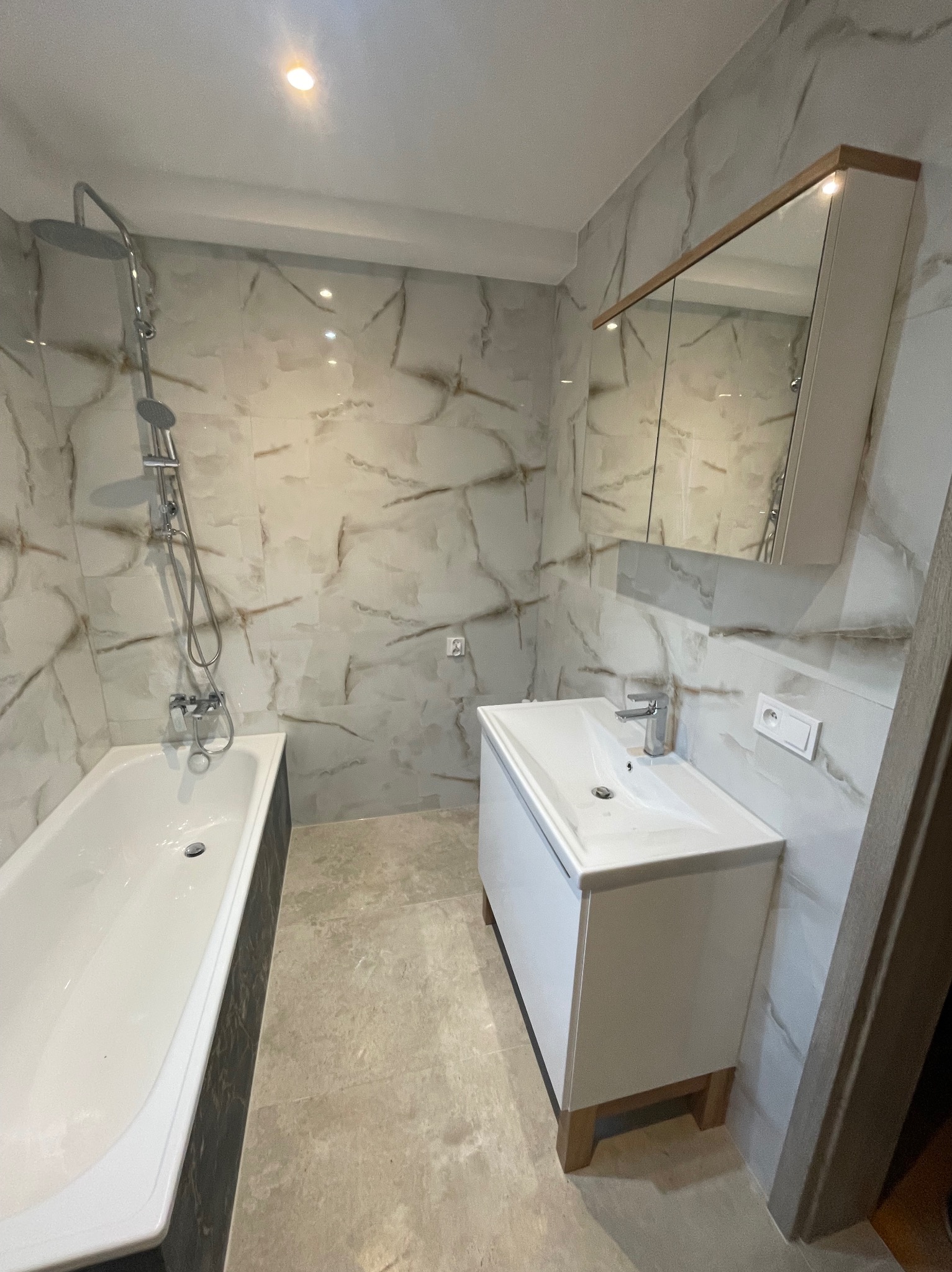 Nowoczesna łazienka po remoncie: biała wanna z czarnym obramowaniem, umywalka z szafką i lustro w drewnianej ramie na tle marmurowych płytek.