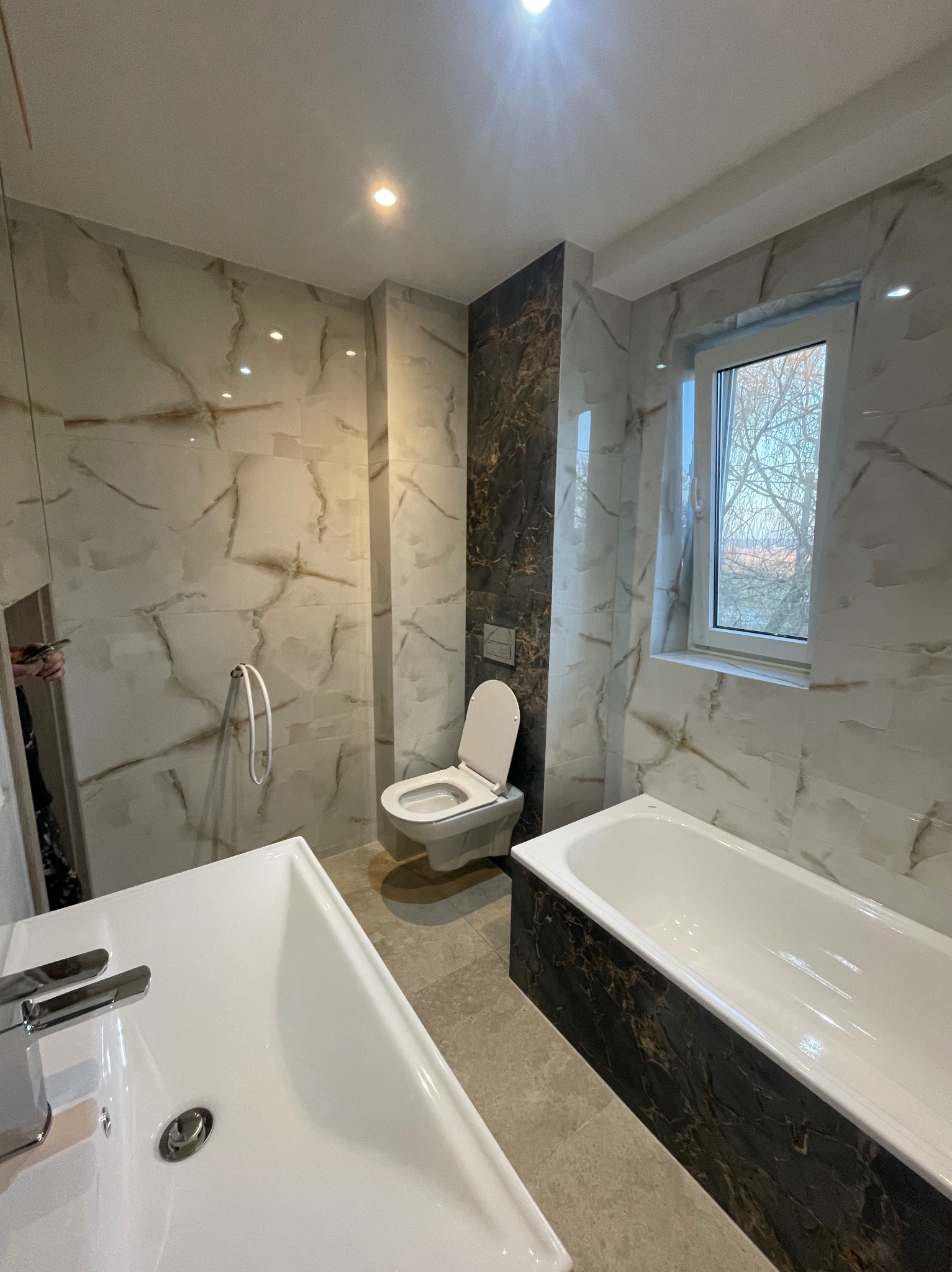 Elegancka łazienka po remoncie: biała armatura, marmurowe płytki z brązowymi żyłami, kabina prysznicowa ze szklaną ścianką i widokiem na zewnątrz przez okno.