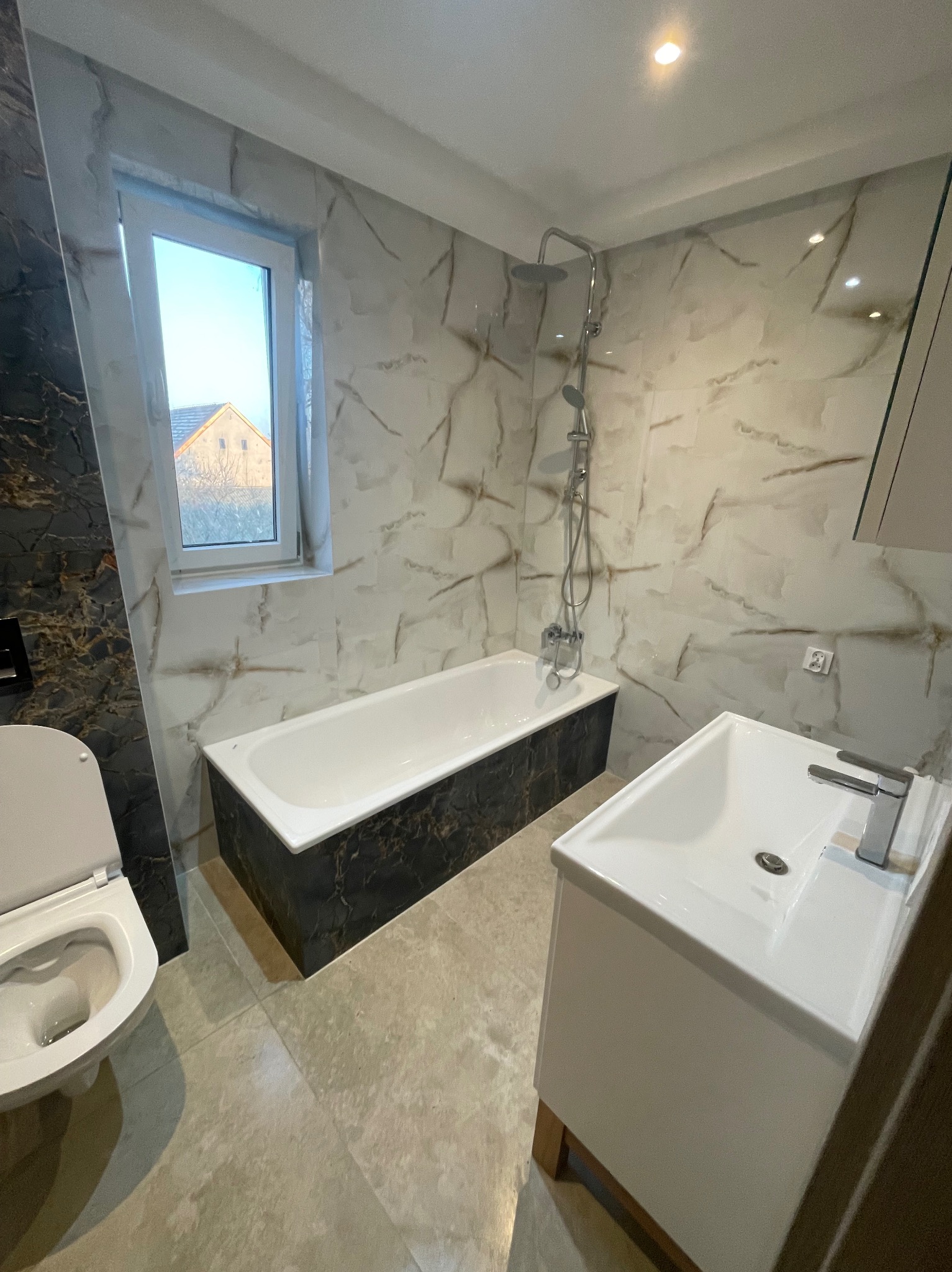Nowoczesna łazienka z marmurowymi płytkami, białą wanną obudowaną czarnym marmurem, umywalką z szafką i toaletą. Widok na okno z widokiem na budynek.