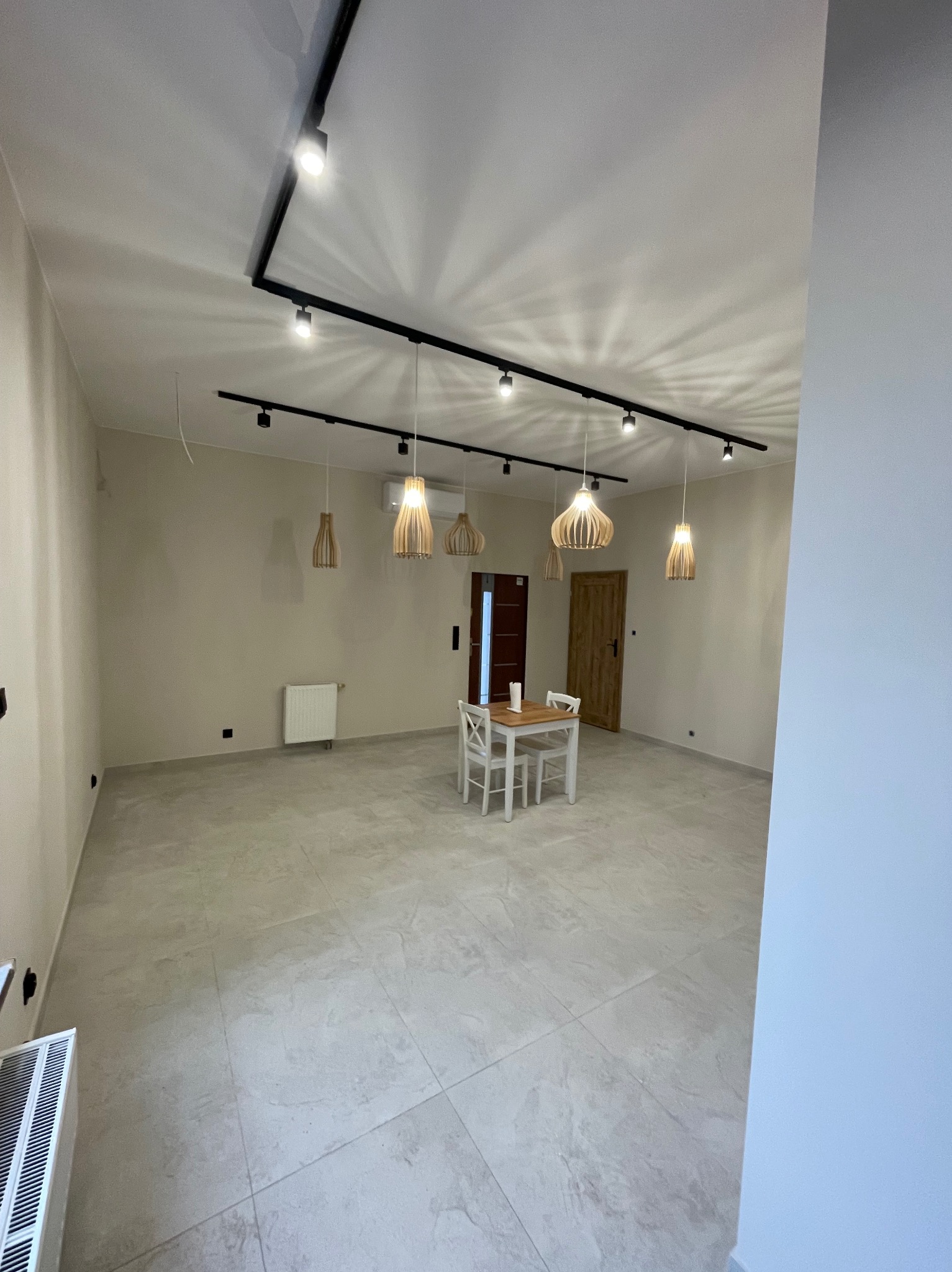 Jasne wnętrze po remoncie z szarą podłogą z płytek, białymi ścianami i drewnianymi lampami wiszącymi. Widoczny stół z krzesłami i grzejnik.