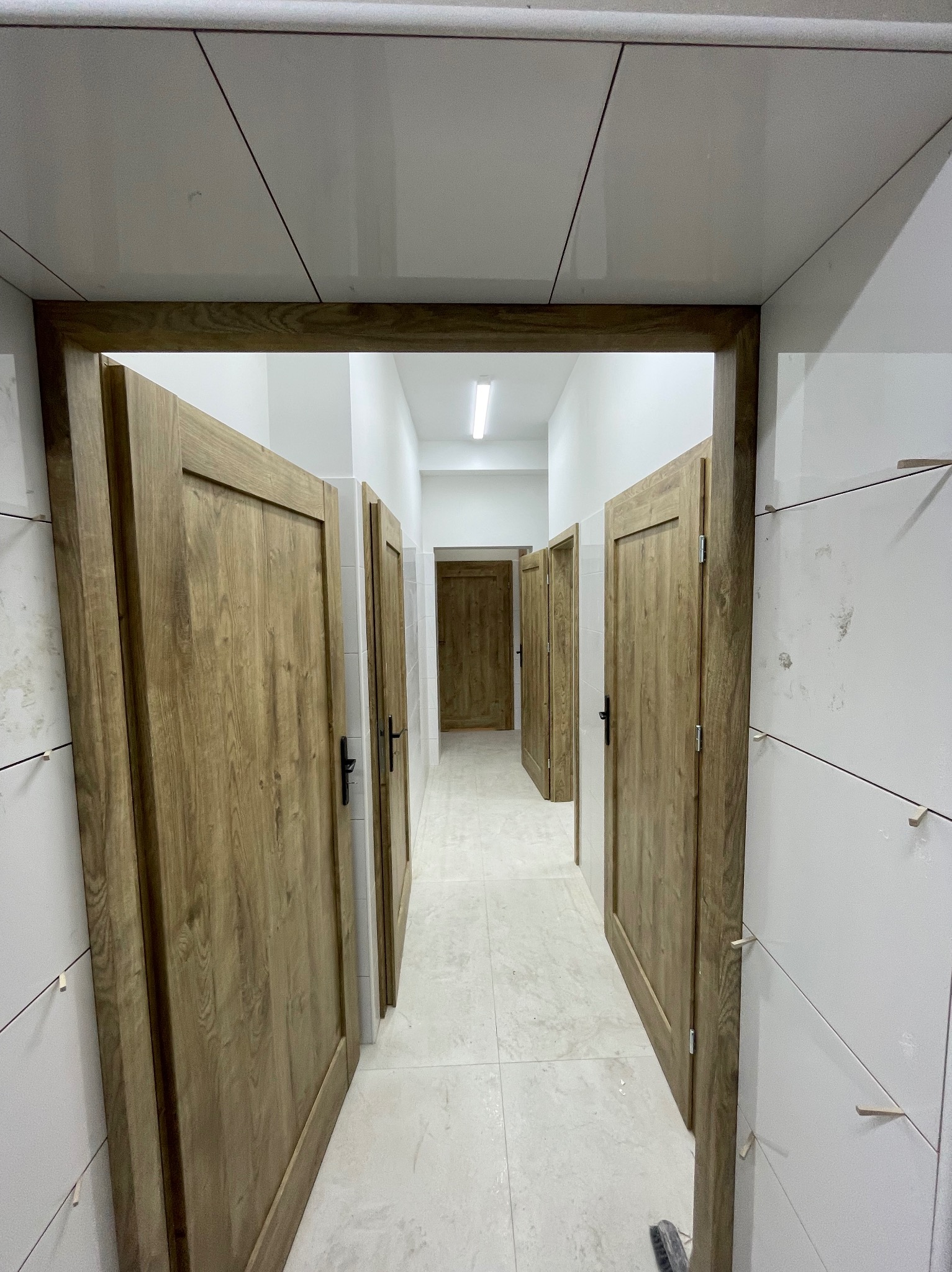Wykończenie łazienki w Raszkowie: widok na korytarz z drewnianymi drzwiami i białymi płytkami, z widocznymi elementami montażowymi na ścianach.