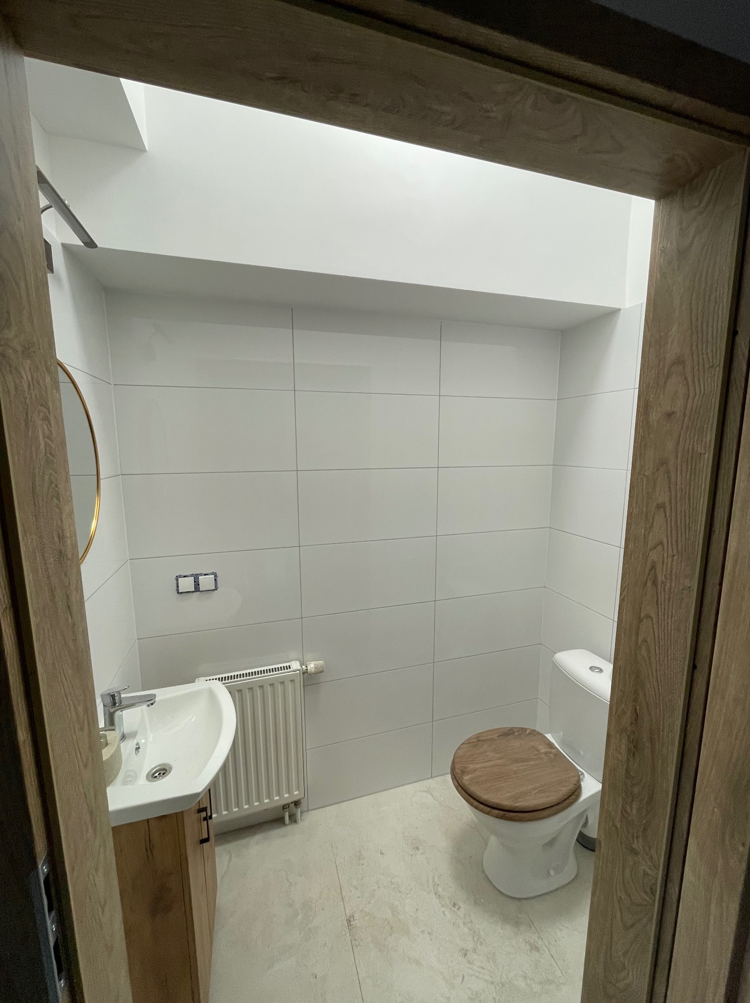 Wykończona łazienka z białymi płytkami, drewnianą deską sedesową i szafką pod umywalką w odcieniach drewna, widziana zza drewnianej futryny drzwi.