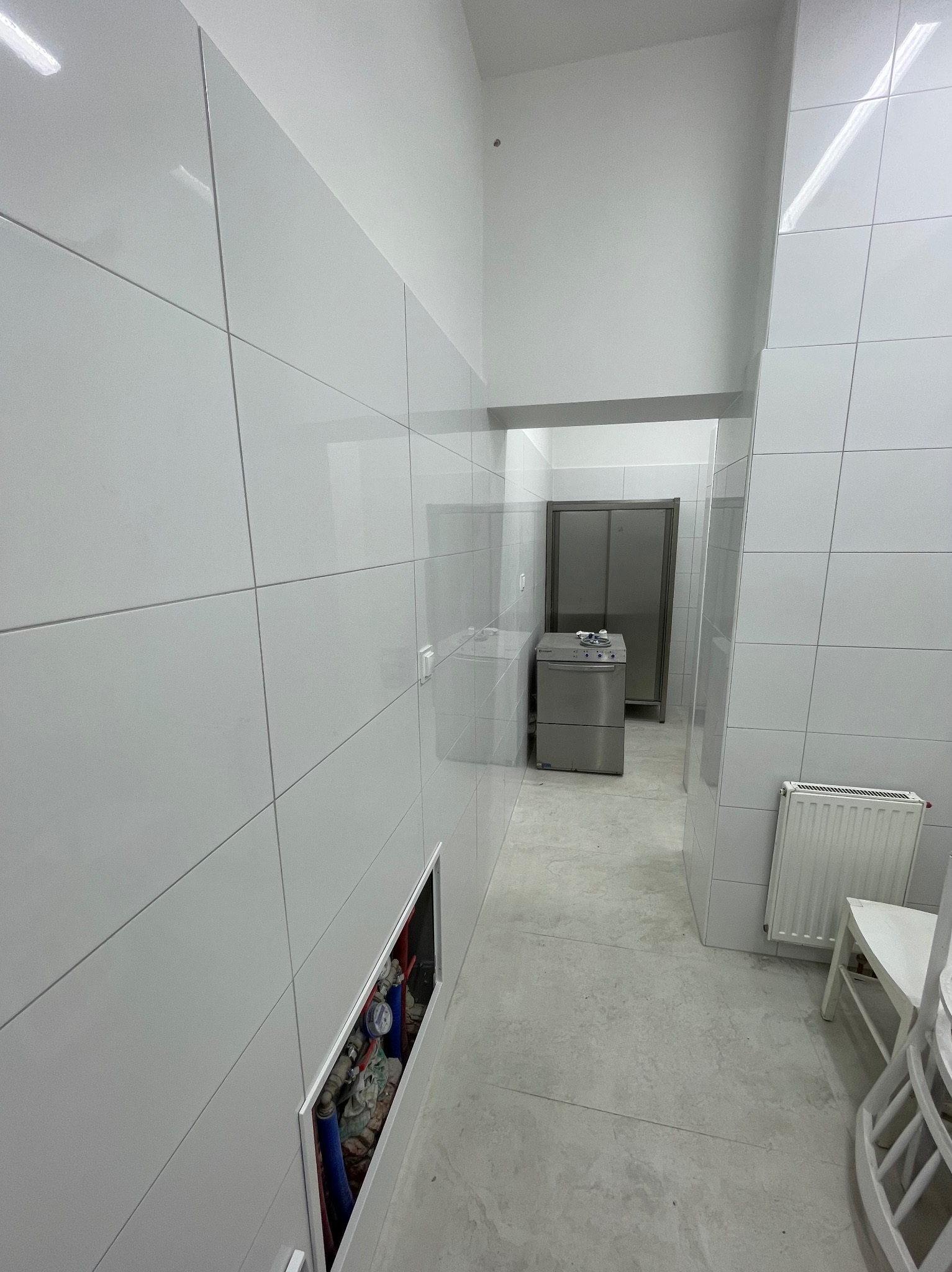 Biała, wykafelkowana ściana z otwartą wnęką instalacyjną, widok na korytarz z metalową szafą i sprzętem. Minimalistyczne wykończenie, surowy styl.