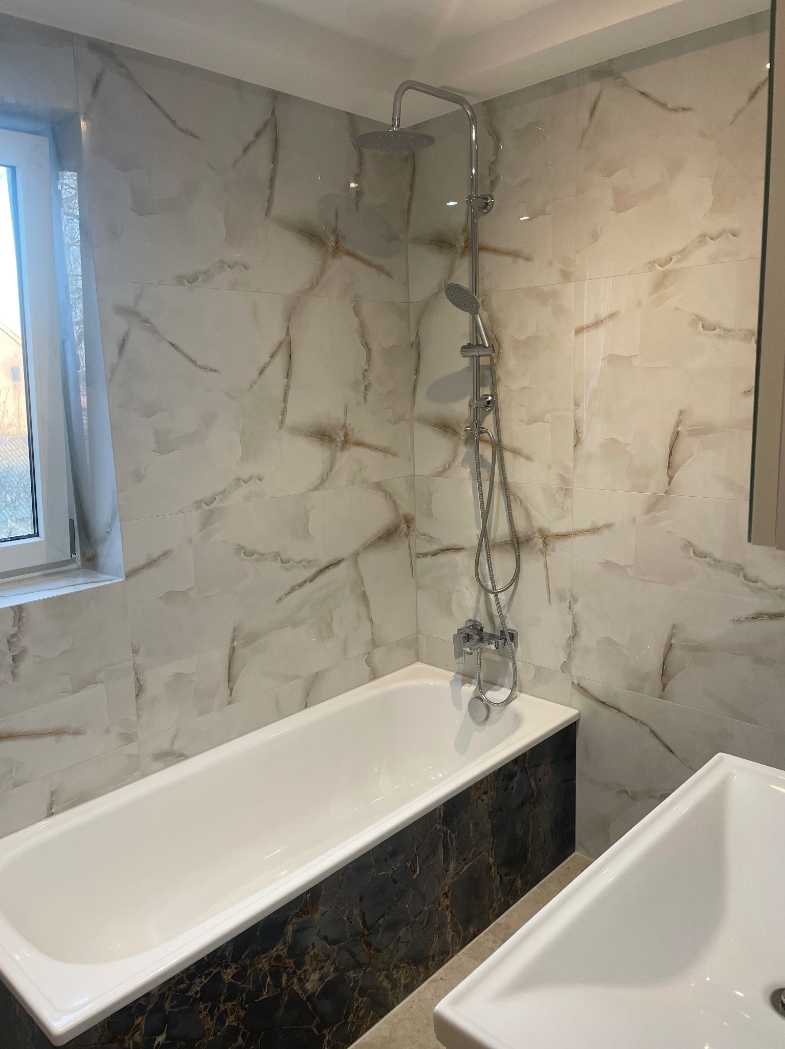 Wykończona łazienka z wanną obudowaną czarnym marmurem, białą umywalką i armaturą w kolorze chromu. Ściany wyłożone płytkami imitującymi marmur.