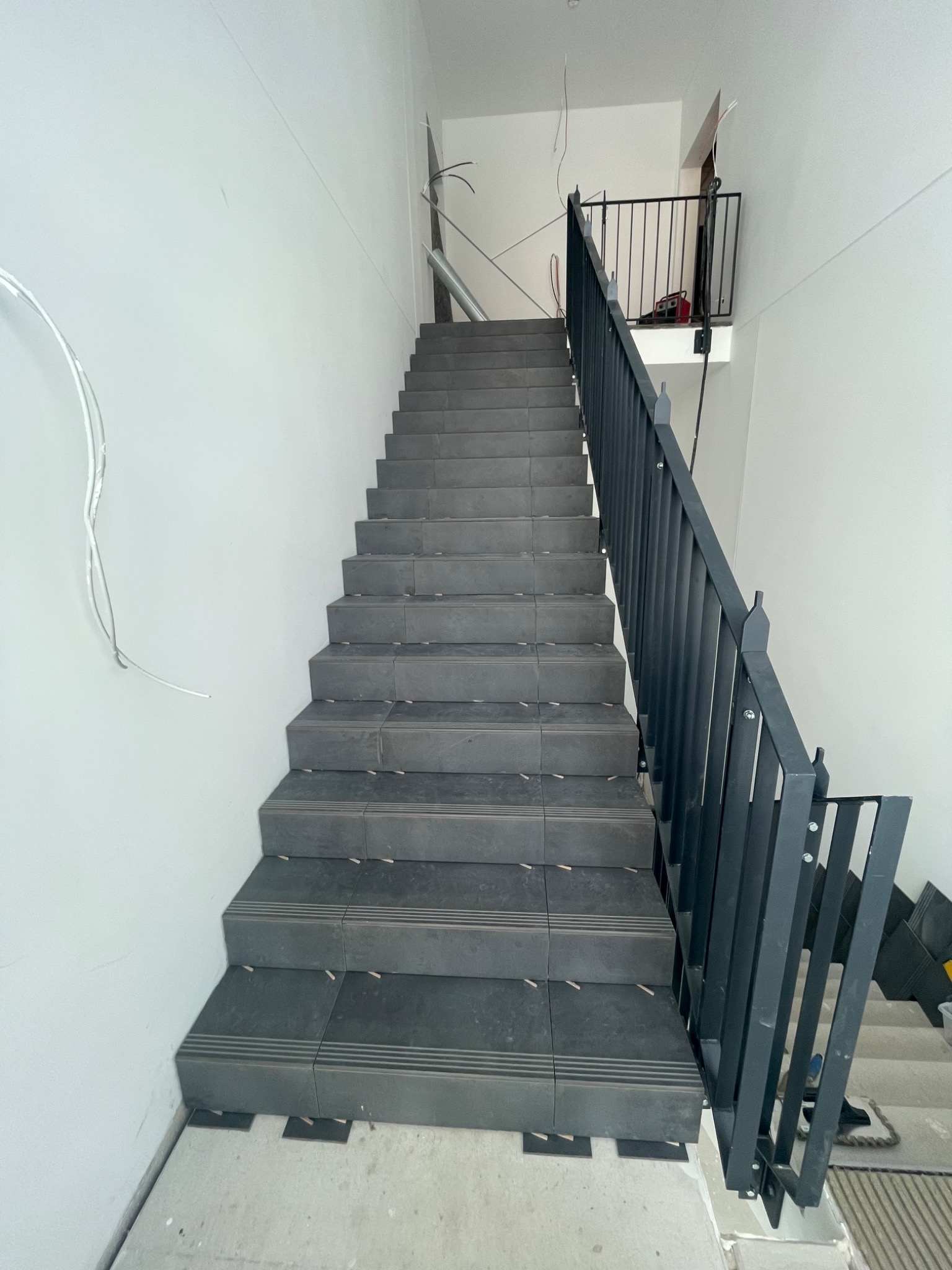 Wykończone schody z ciemnej terakoty z czarną balustradą w jasnym wnętrzu. Widoczne elementy instalacji elektrycznej i płytki na podłodze.