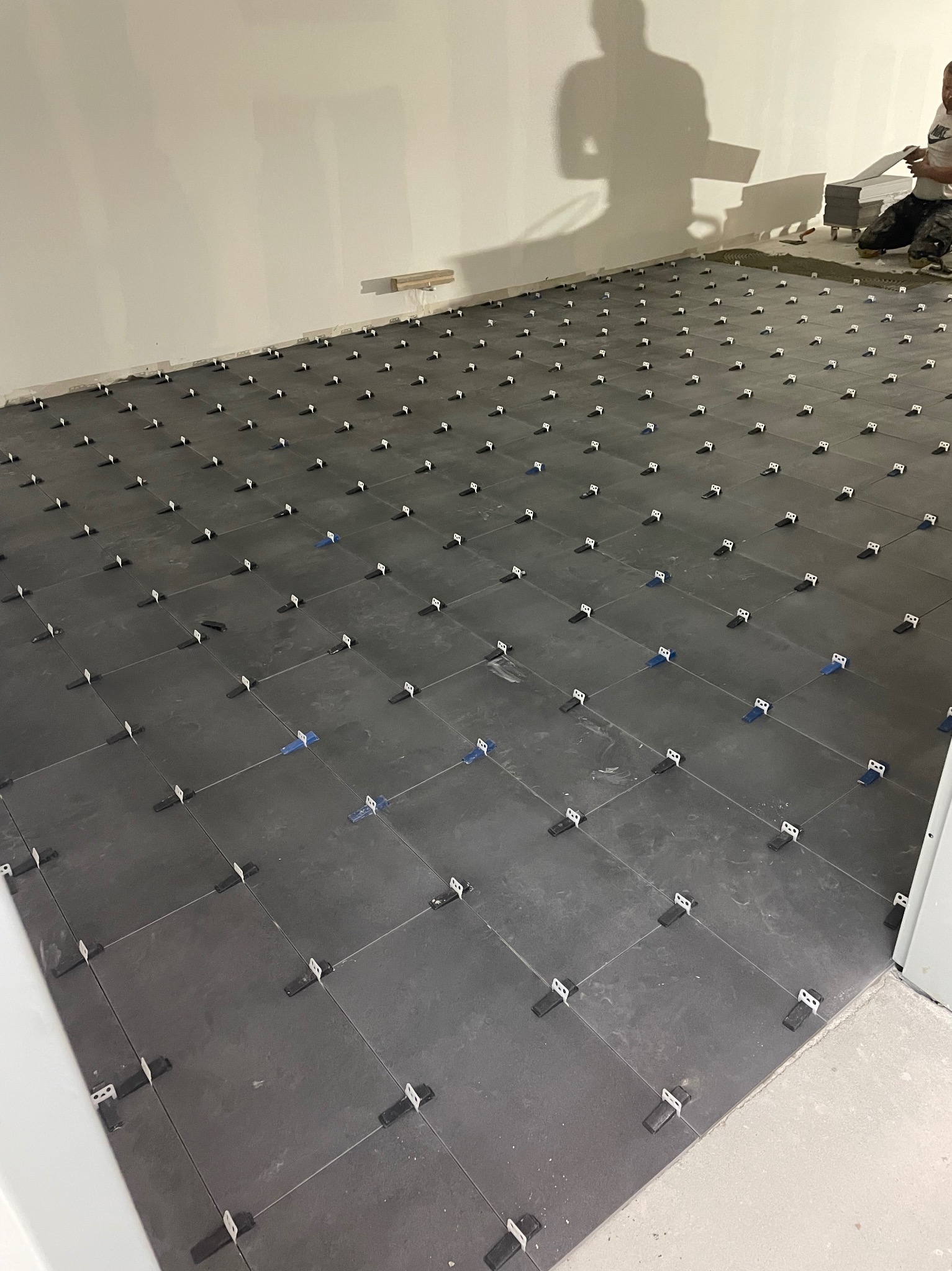 Ciemnoszare płytki podłogowe, częściowo ułożone, z systemem poziomowania płytek. W tle cień osoby układającej płytki. Widok z góry, perspektywa.