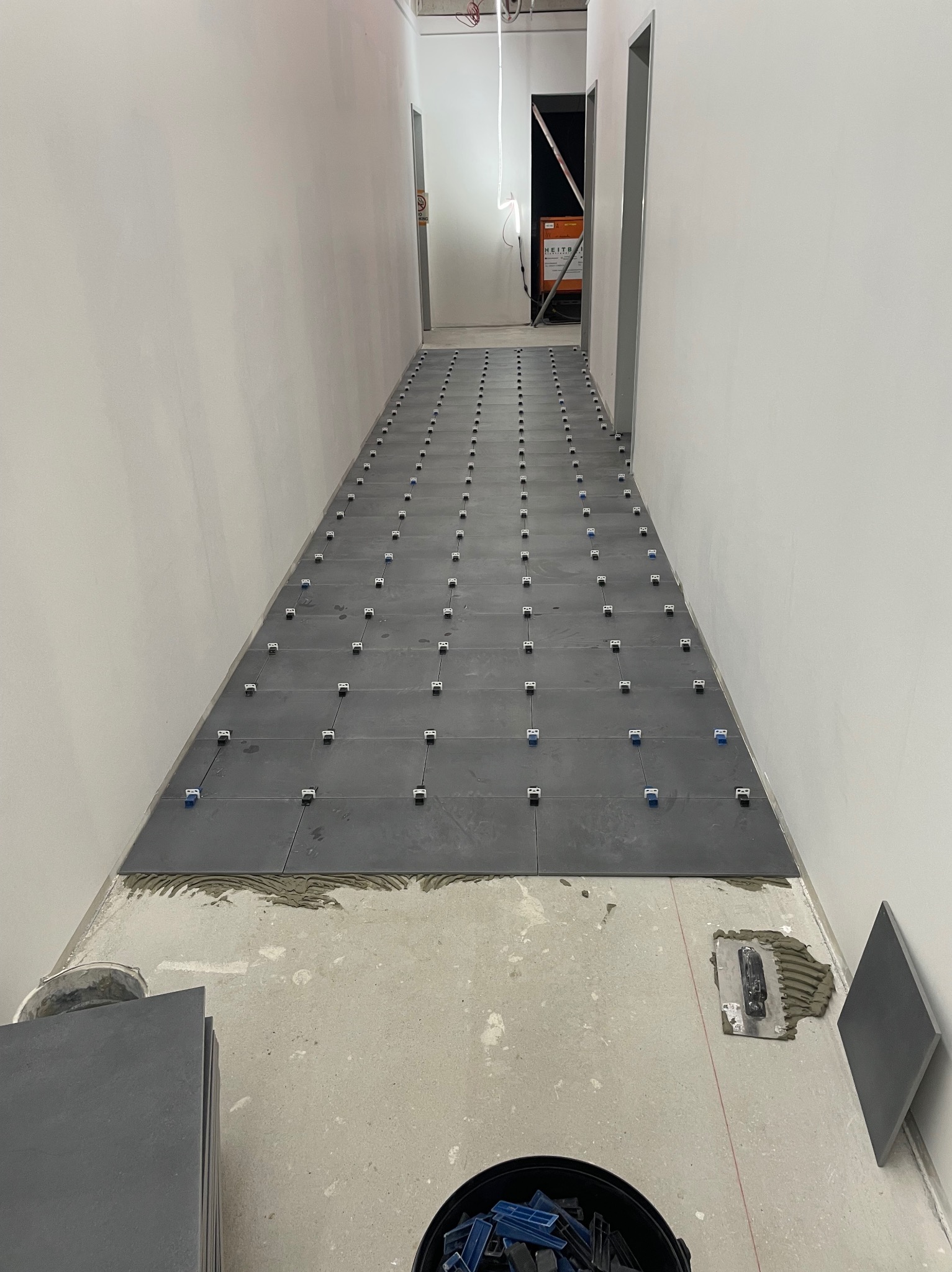 Układanie ciemnoszarych płytek podłogowych w korytarzu, z użyciem klipsów poziomujących. Widoczne narzędzia glazurnicze i zapas płytek. Surowe ściany w tle.