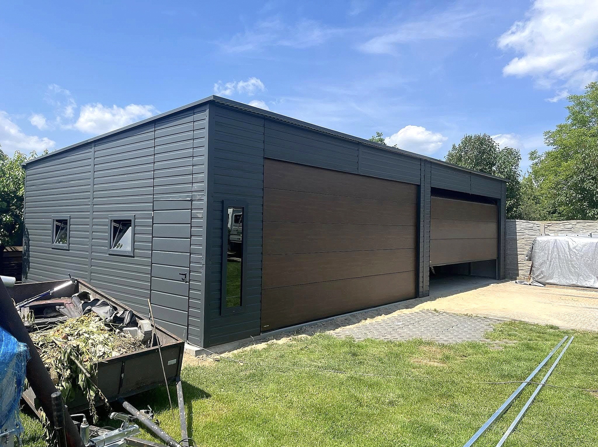 Nowoczesny garaż blaszany z brązową bramą segmentową i szarymi ścianami, widoczny z zewnątrz na tle zielonego trawnika i błękitnego nieba.