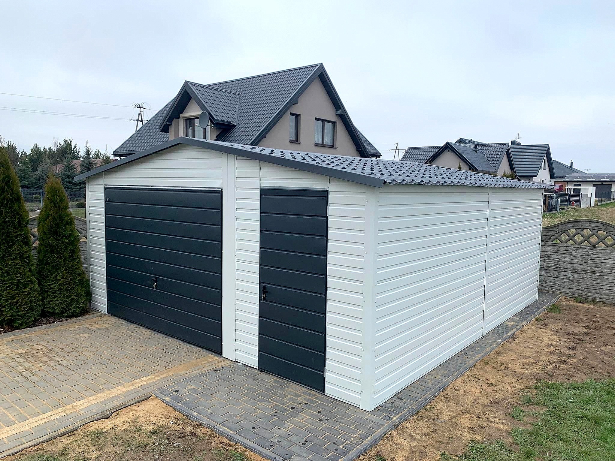 Biało-grafitowy garaż blaszany z dwoma bramami i drzwiami wejściowymi, na kostce brukowej, z domami w tle. Widok z przodu, pochmurny dzień.