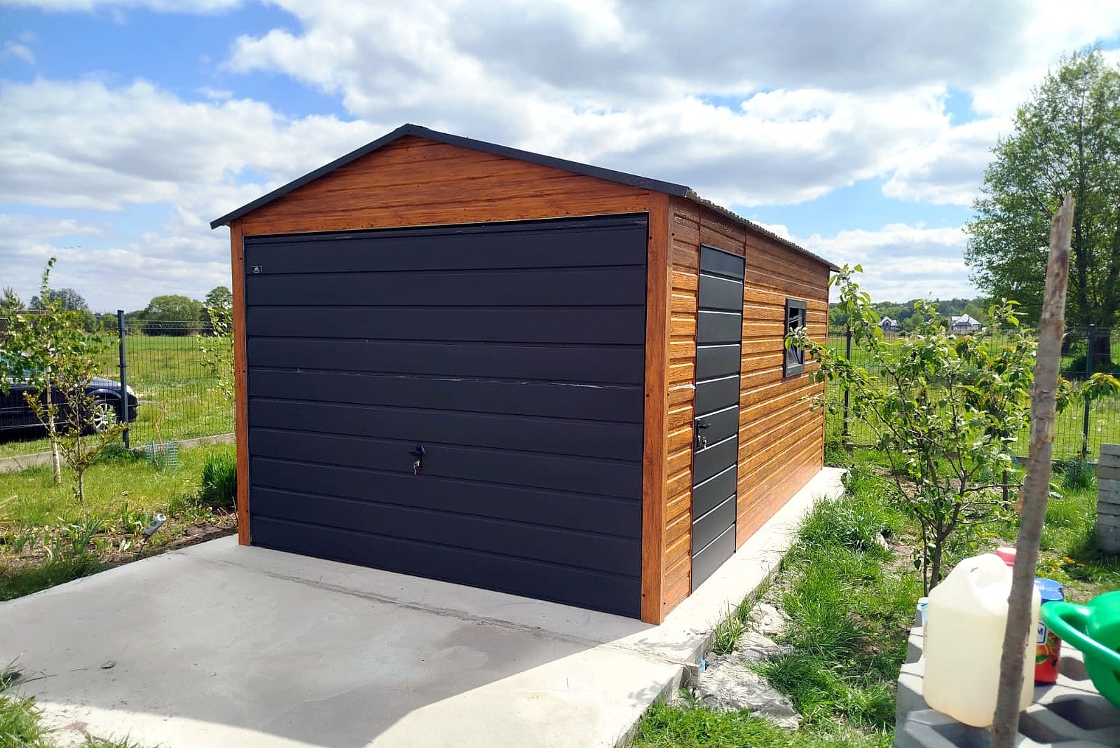 Gotowy garaż blaszany z drewnopodobną elewacją i antracytową bramą segmentową, usytuowany na betonowej wylewce, w tle zielony krajobraz.