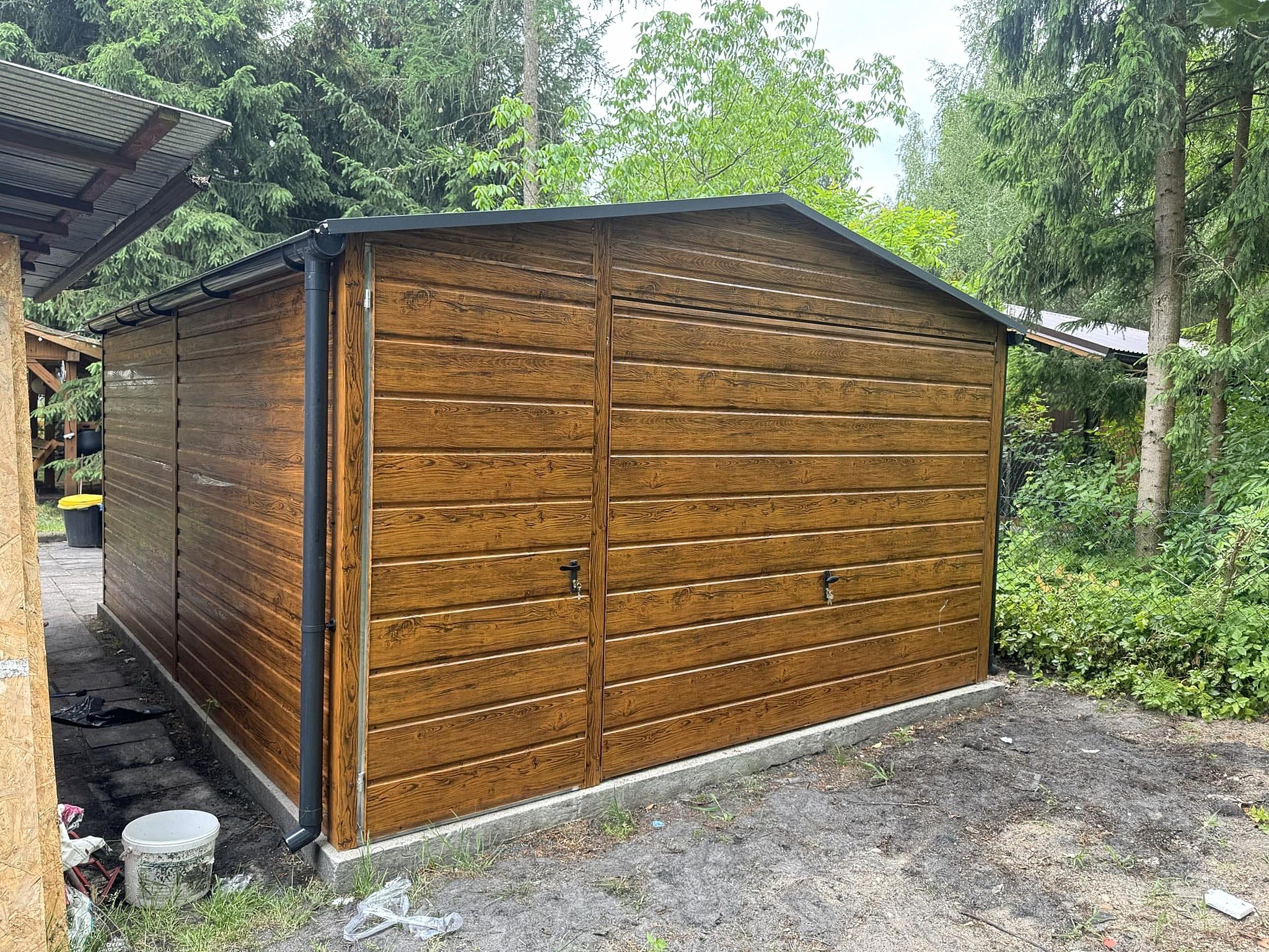 Garaż blaszany z drewnopodobną elewacją, dwuspadowy dach z czarną obróbką blacharską, widoczne rynny, otoczenie zieleni, częściowo widoczny teren.