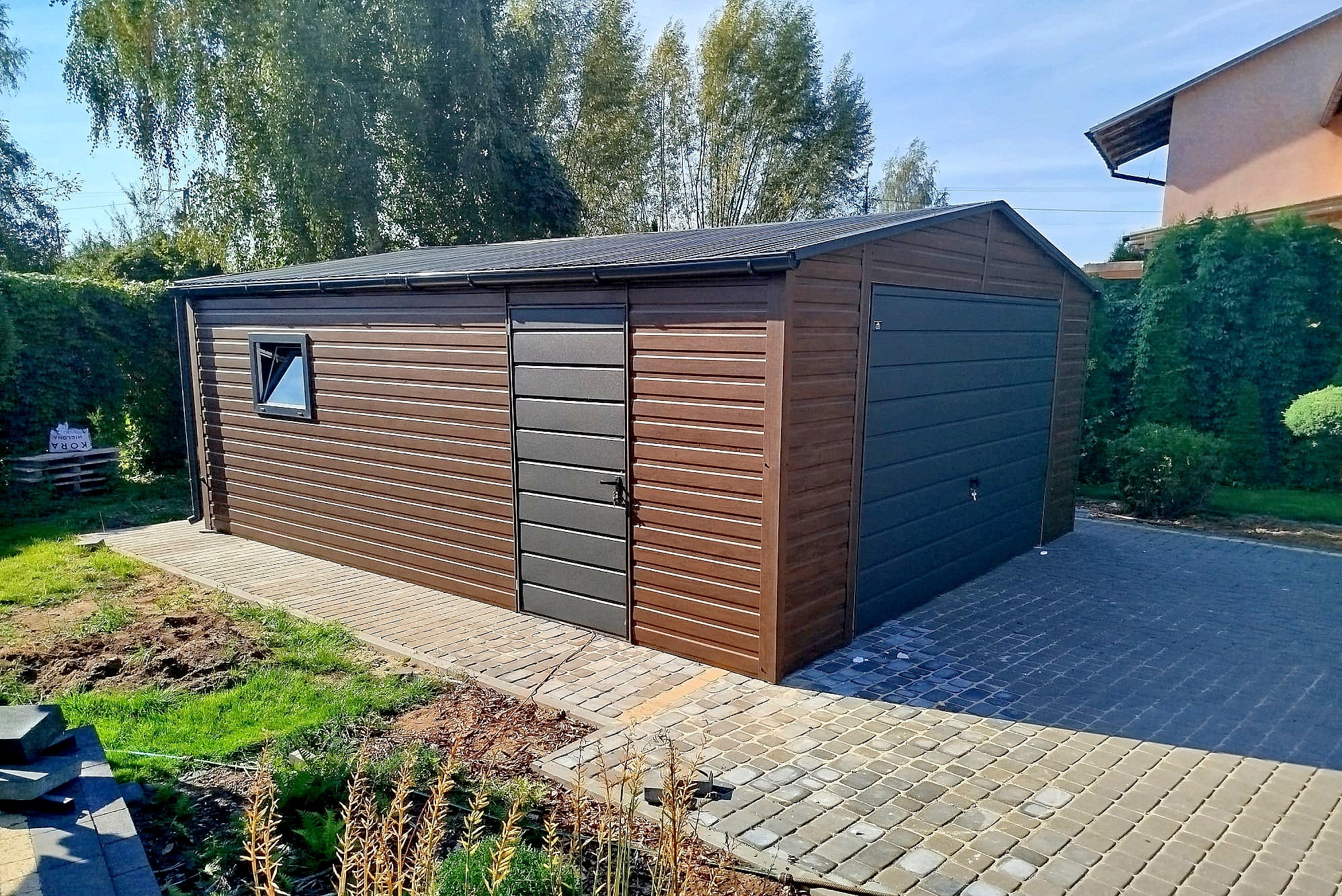 Brązowy garaż blaszany z szarą bramą i drzwiami, otoczony kostką brukową i zielenią. Widoczne okno i elementy wykończeniowe. Garaż na tle drzew i domu.
