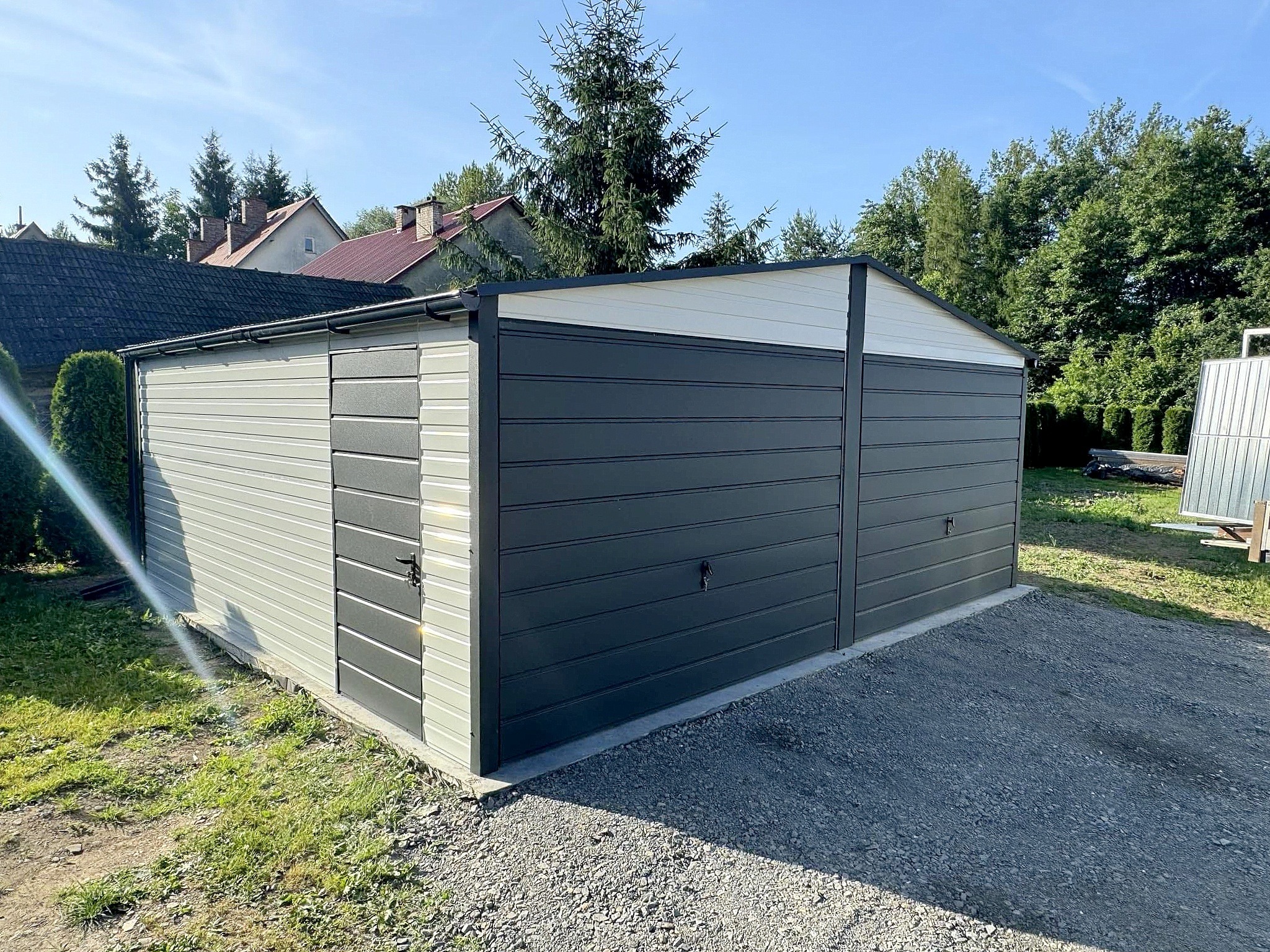 Dwukolorowy garaż blaszany z dwiema bramami, na żwirowym podłożu, w tle zieleń i zabudowania. Garaż z blachy trapezowej, z widocznym systemem odprowadzania wody.