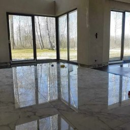 Od remontu do efektu - remonty i wykończenia wnętrz - Błyszcząca, marmurowa podłoga w jasnym wnętrzu z dużymi oknami, przez które widać las. Częściowo wykończone ściany i widoczne narzędzia sugerują etap prac wykończeniowych.